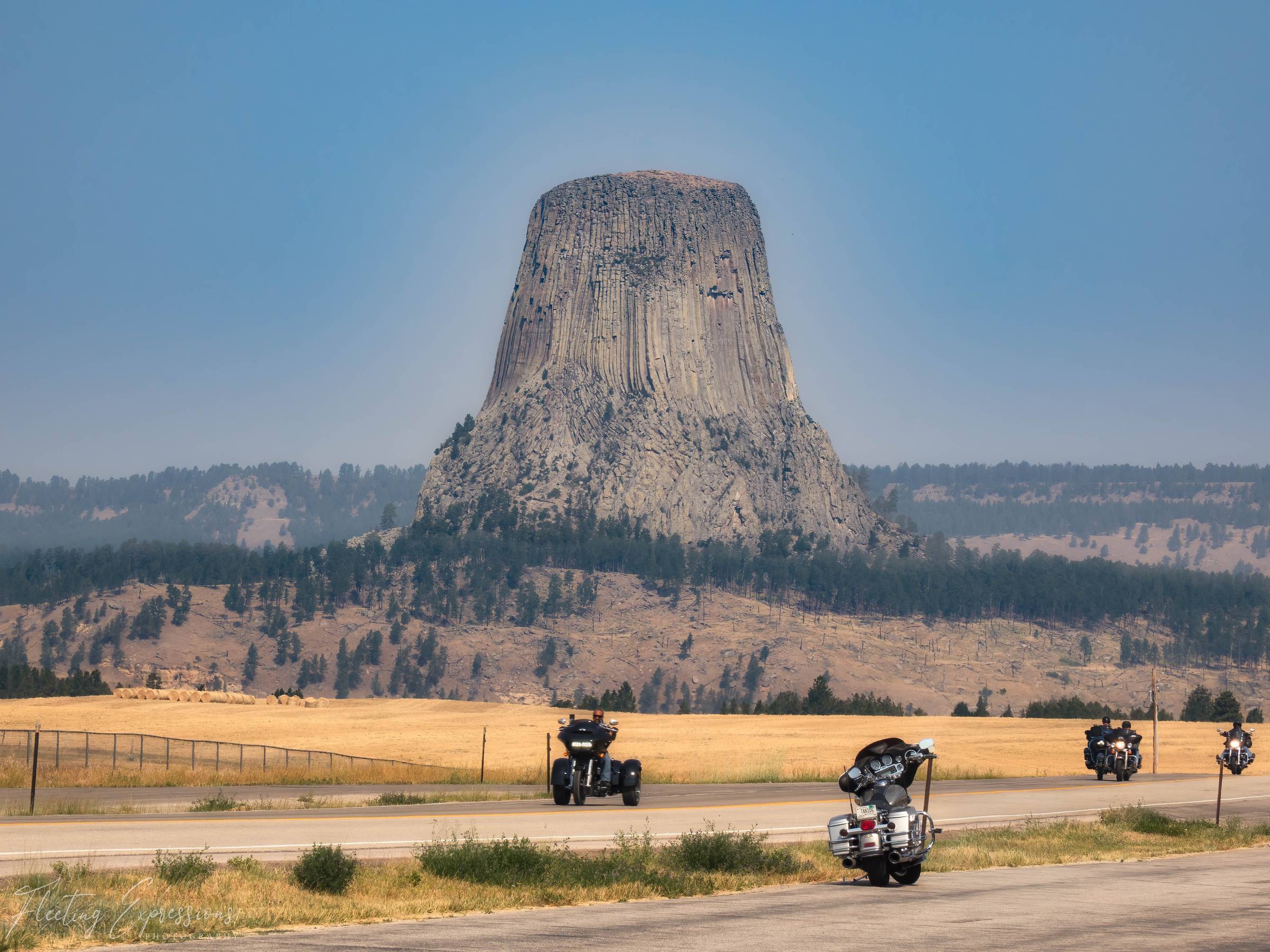 Devils Tower rising above a grassy field with a group of motorcycles and riders on the road in the foreground, hazy blue sky and pine-covered hills in the distance. 