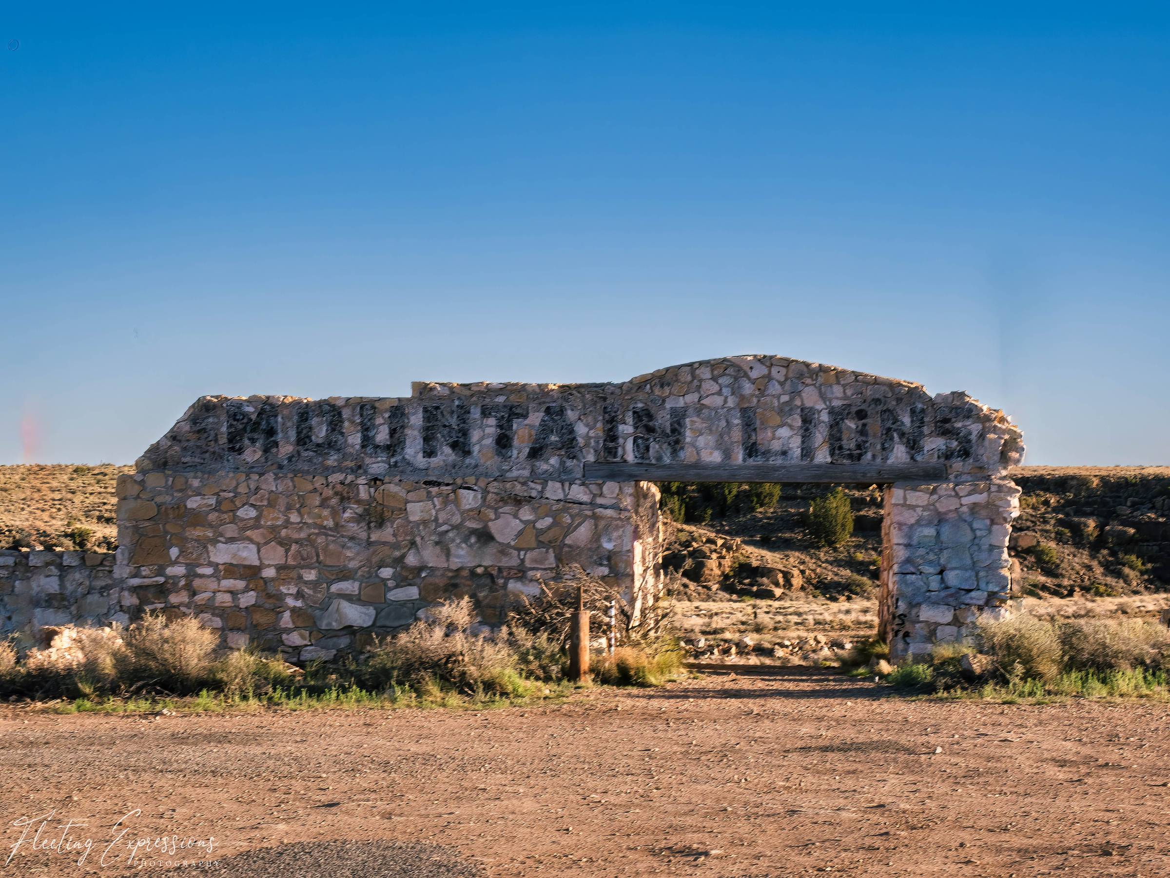 A crumbling stone wall with the words Mountain Lions carved into it stands against a vivid blue sky at the abandoned Two Guns ghost town along Arizona Route 66.