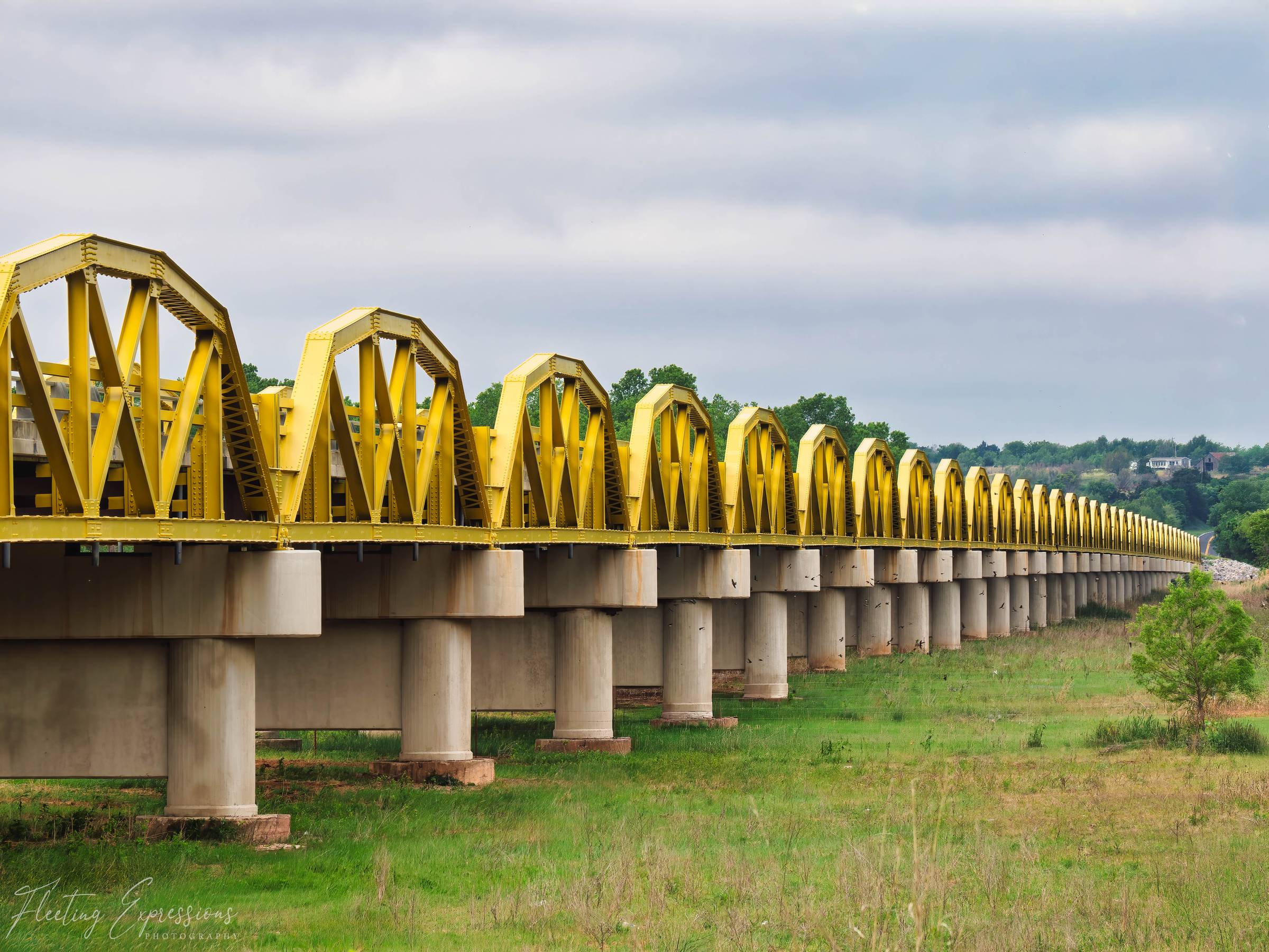 A long yellow pony truss bridge stretches across a flat green landscape over the South Canadian River near Bridgeport, Oklahoma, its repeating steel arches receding into the distance.