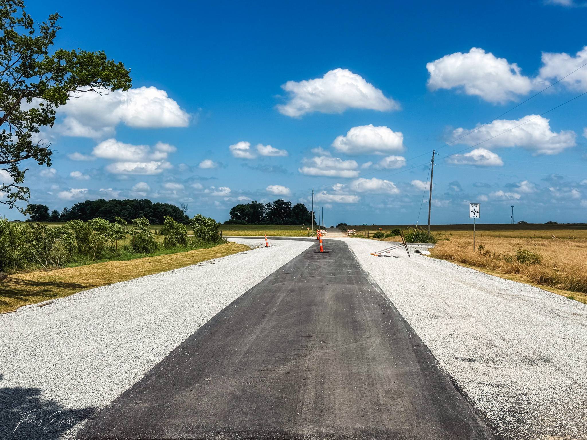 A narrow strip of dark pavement runs straight through green and gold fields under a wide sky with scattered clouds.