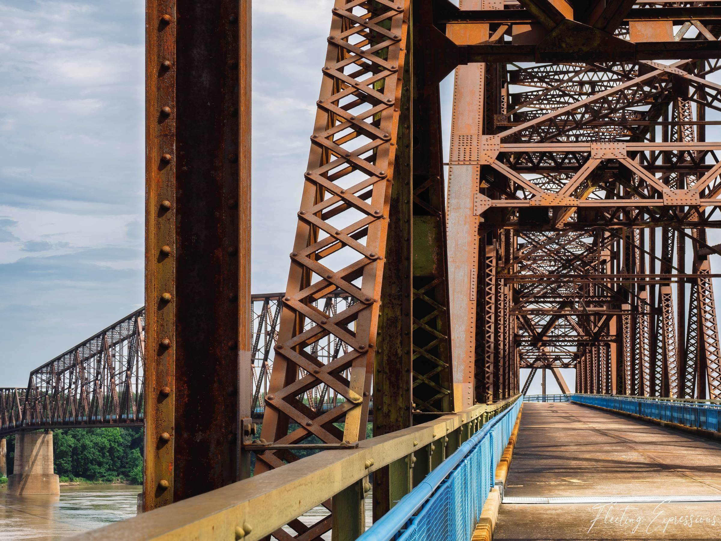 Steel trusswork of the Old Chain of Rocks Bridge stretches into the distance, its deck framed by riveted diagonals; a second historic bridge is visible in the left background above the Mississippi River.