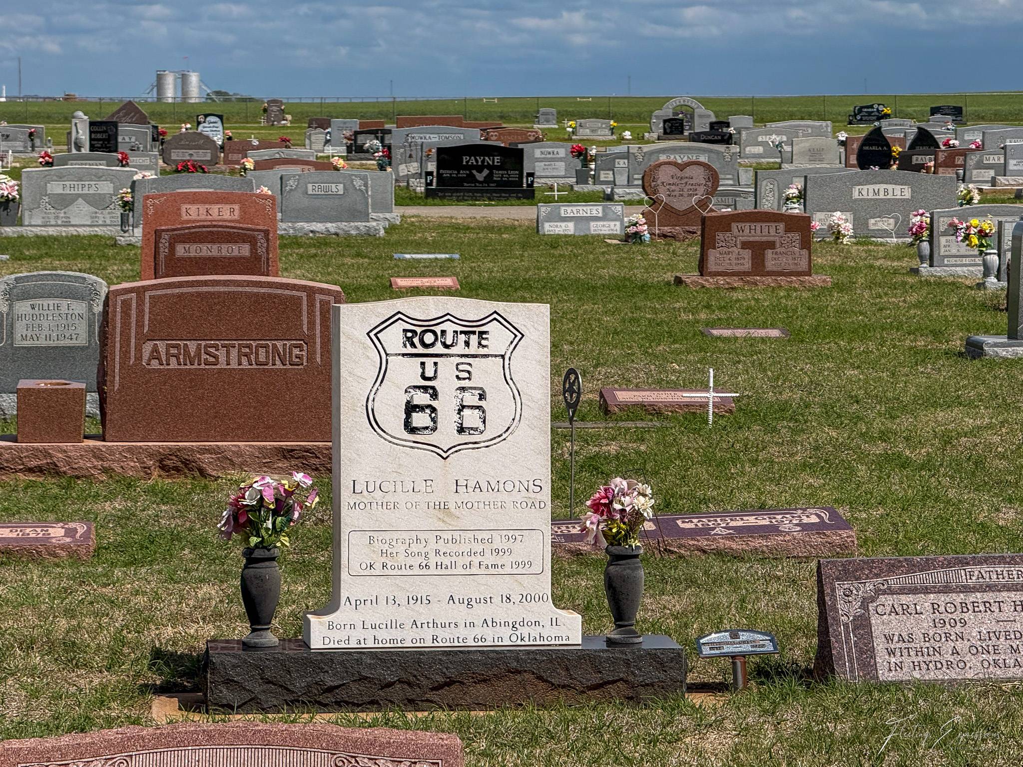 A white marble headstone engraved with a Route 66 shield marks the grave of Lucille Hamons, "Mother of the Mother Road," in a small Oklahoma cemetery. The inscription reads "Died at home on Route 66 in Oklahoma." Plastic flower arrangements flank the stone, with flat green fields visible in the background.