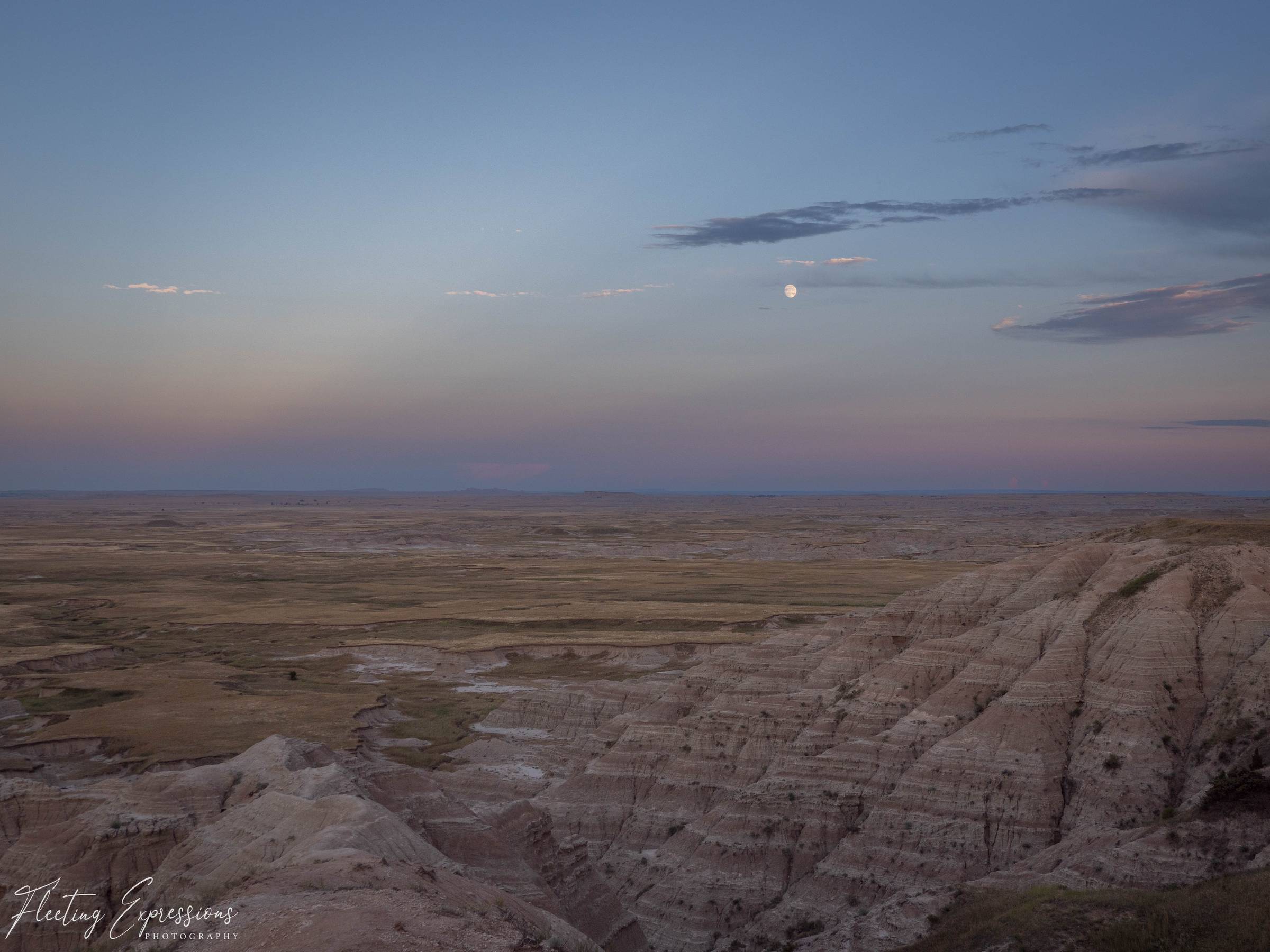 Full moon rising above Badlands rock formations in a fading blue and pink evening sky. 