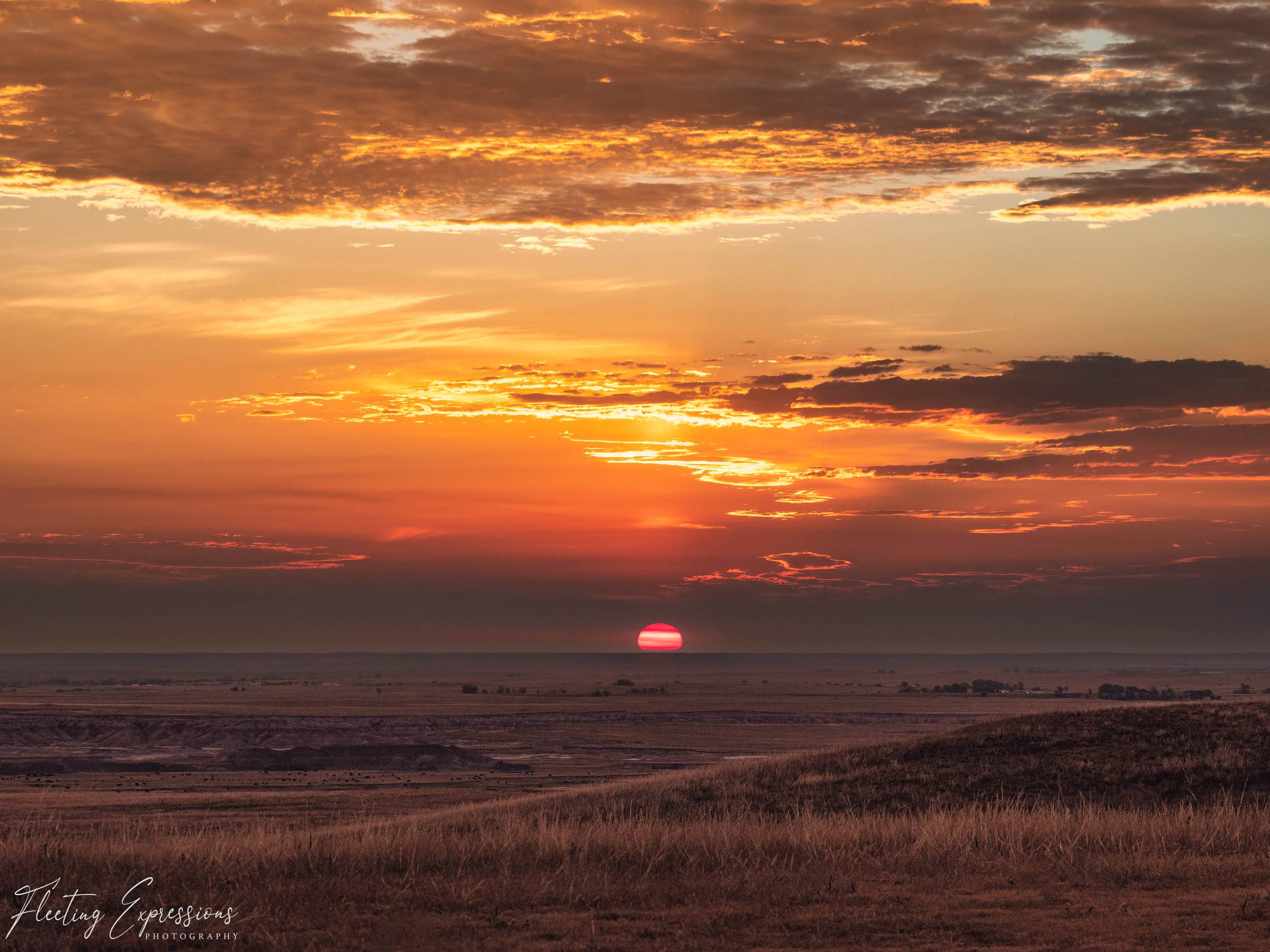 Sun setting low over the Badlands, glowing red through bands of orange cloud above a shadowed grassland valley. 