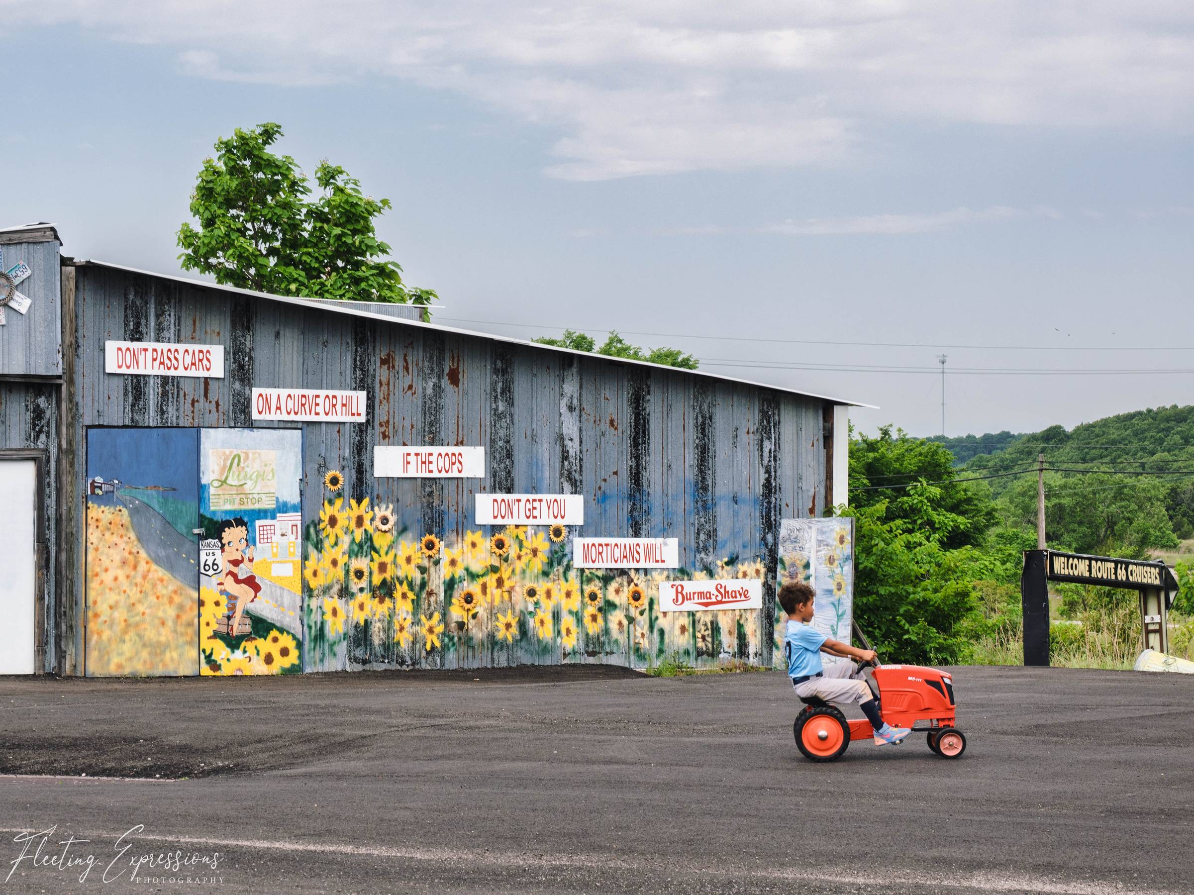 A child pedals a toy tractor past a metal building painted with sunflowers and red Burma-Shave road-safety signs.