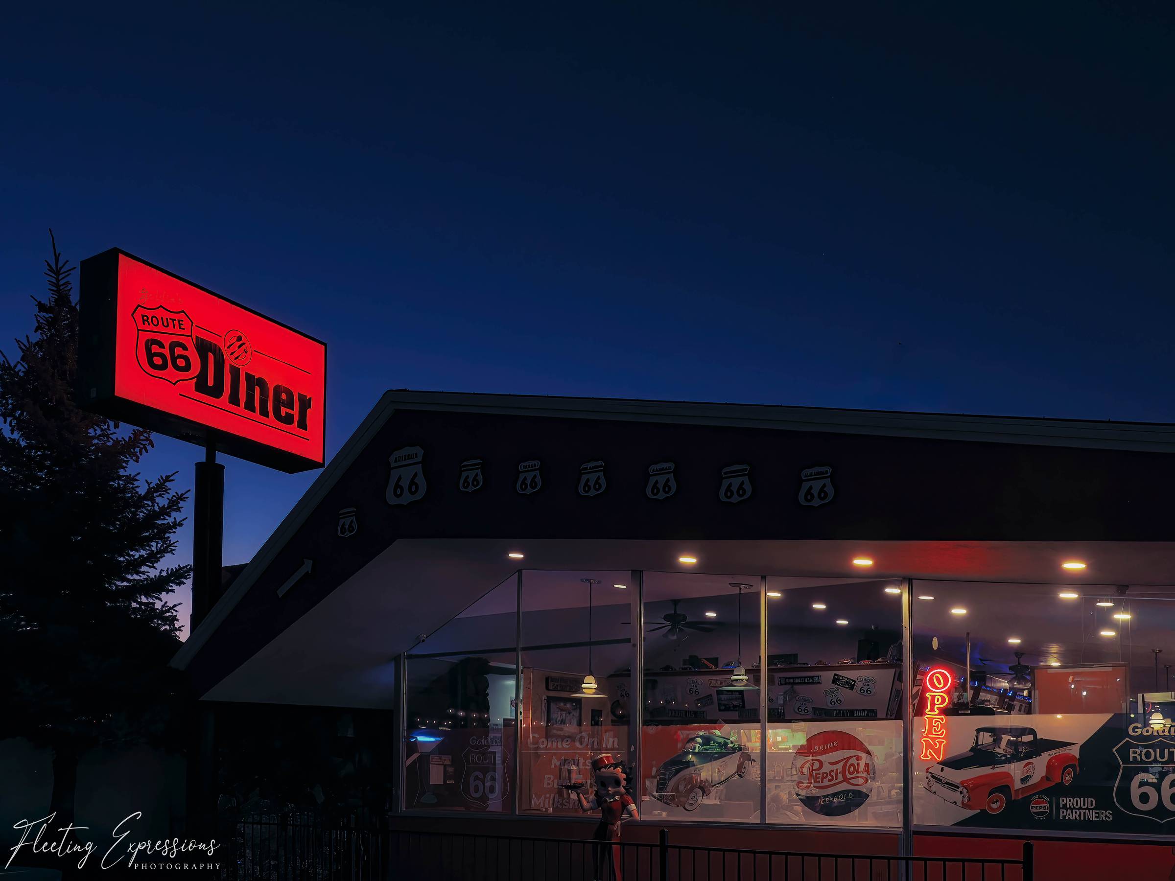 Red neon Route 66 diner sign glowing above a vintage-style diner at dusk, with lit windows, Route 66 decor, and a dark blue evening sky.