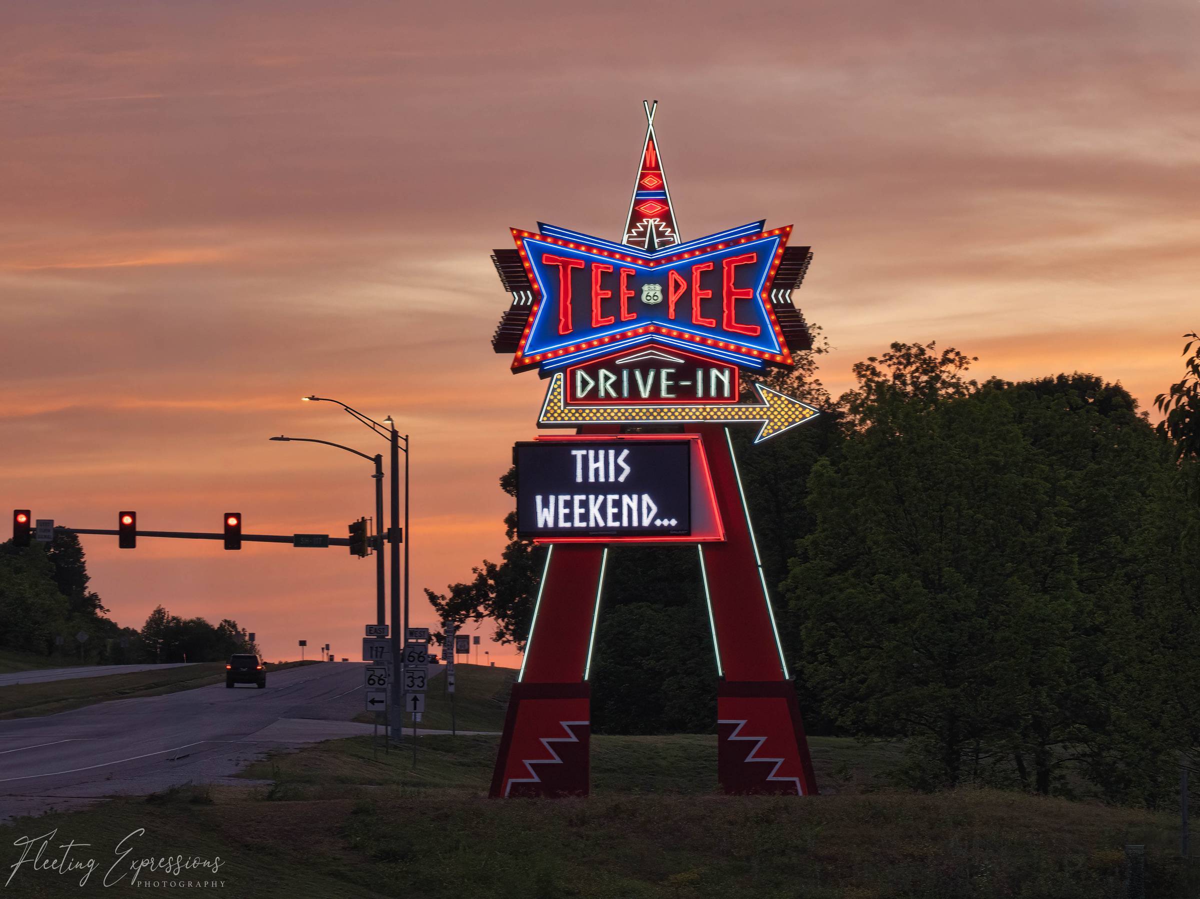 Tall neon Tee Pee Drive-In sign lit up at dusk, shaped like a star and arrow with a reader board that says "This Weekend," against an orange sky.