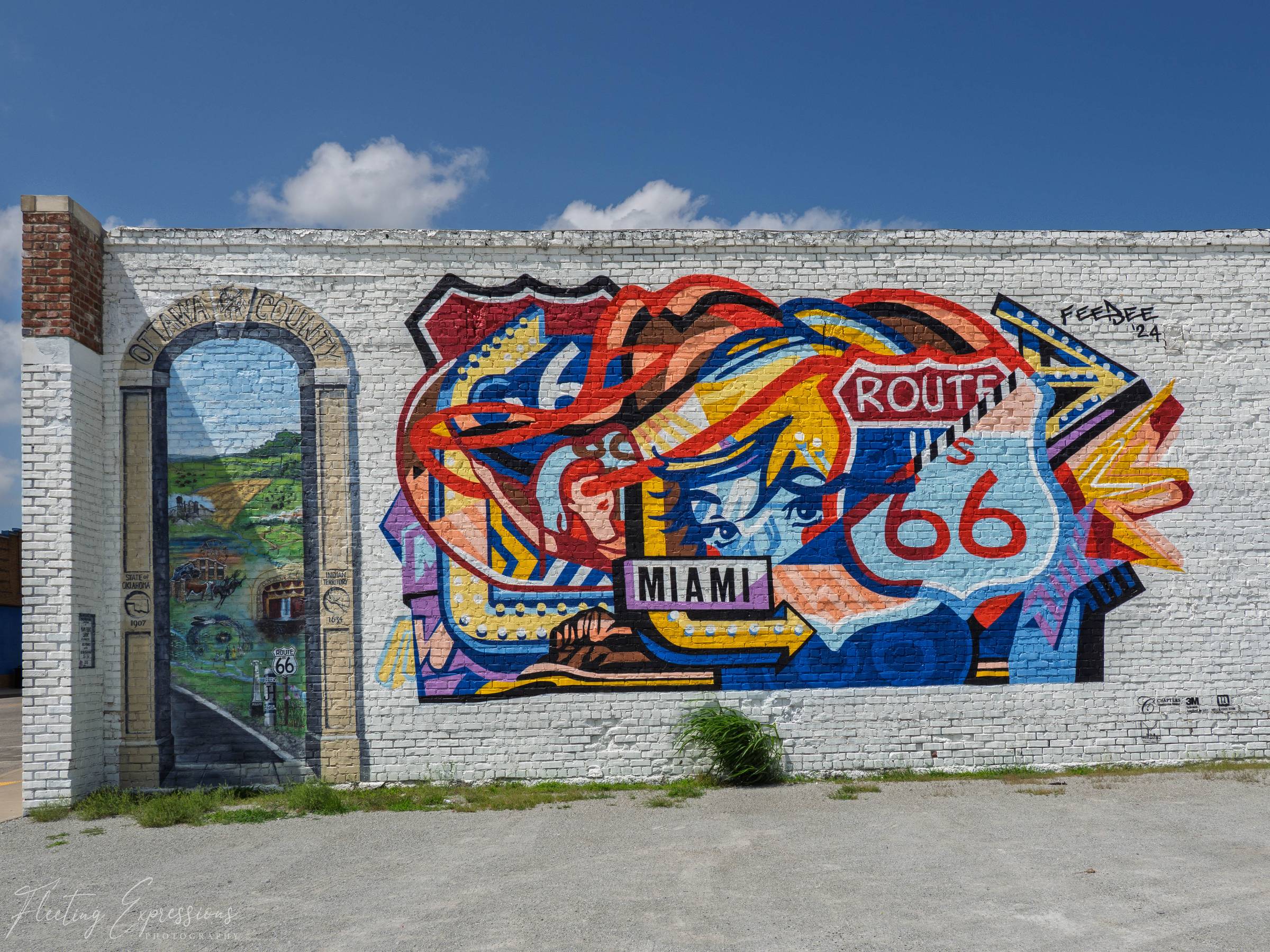 Large, colorful mural painted on a white brick building in Miami, Oklahoma. The artwork features a stylized face, a Route 66 highway shield, bold arrows, and a painted Ottawa County archway framing a rural Route 66 scene under a blue sky.