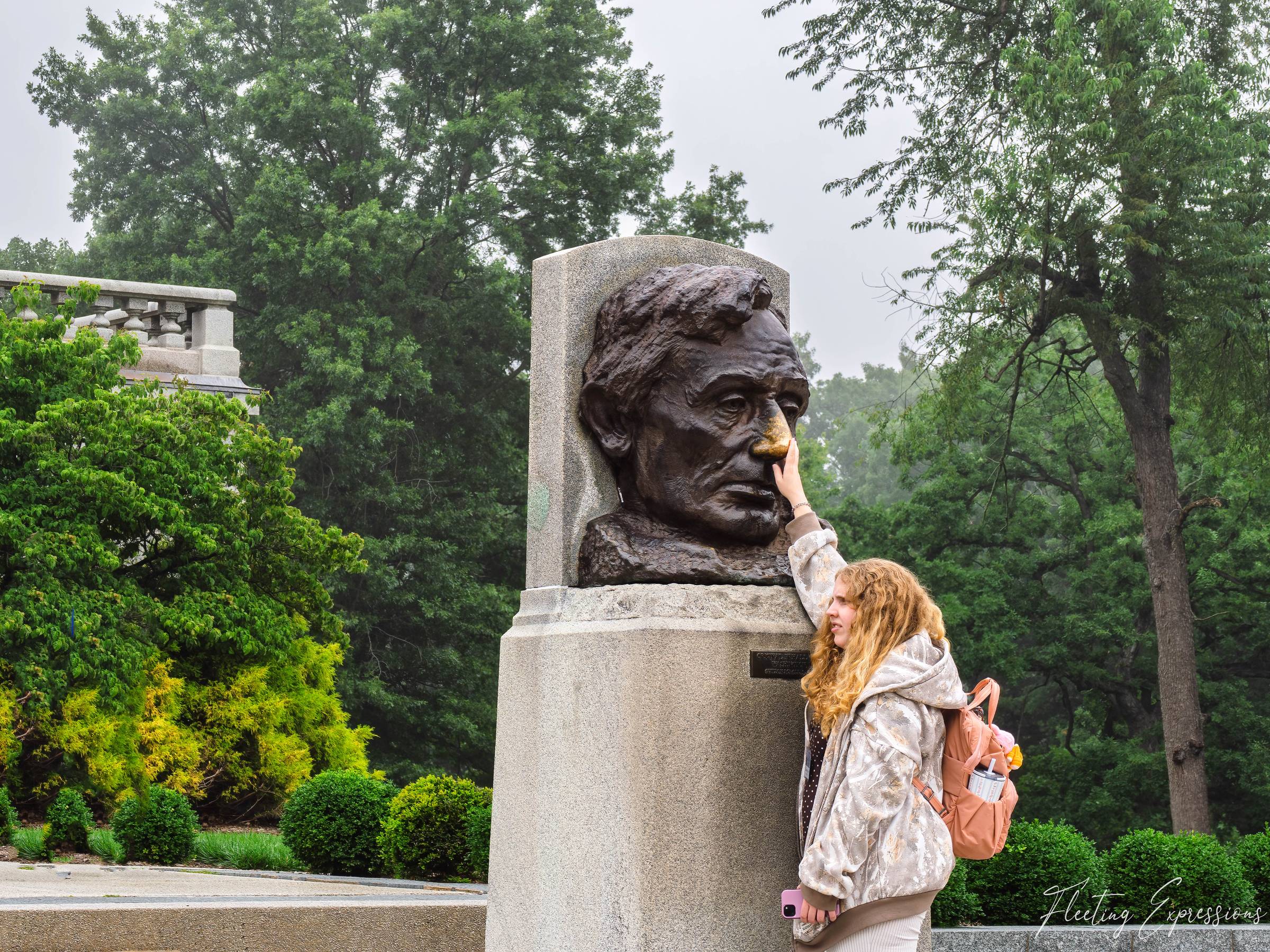 A young woman with curly blonde hair reaches up to touch the golden, polished nose of a bronze Abraham Lincoln bust on a granite pedestal.