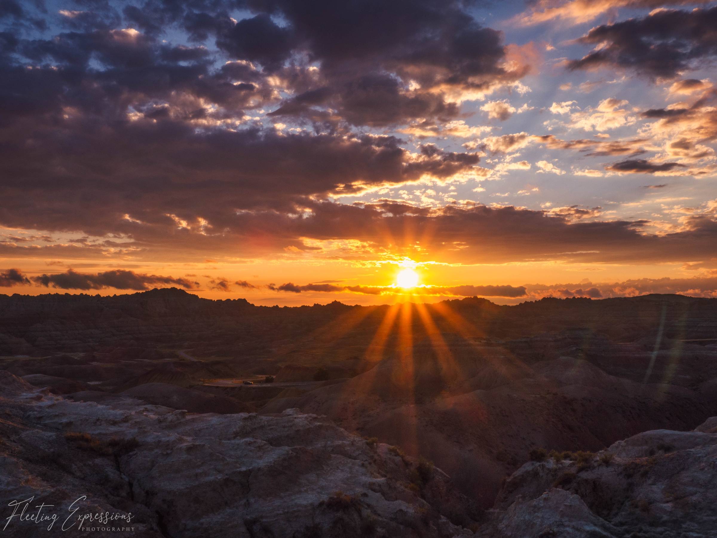 Sun bursting through a silhouetted badlands ridgeline at sunset, orange light rays spreading across a cloud filled sky.