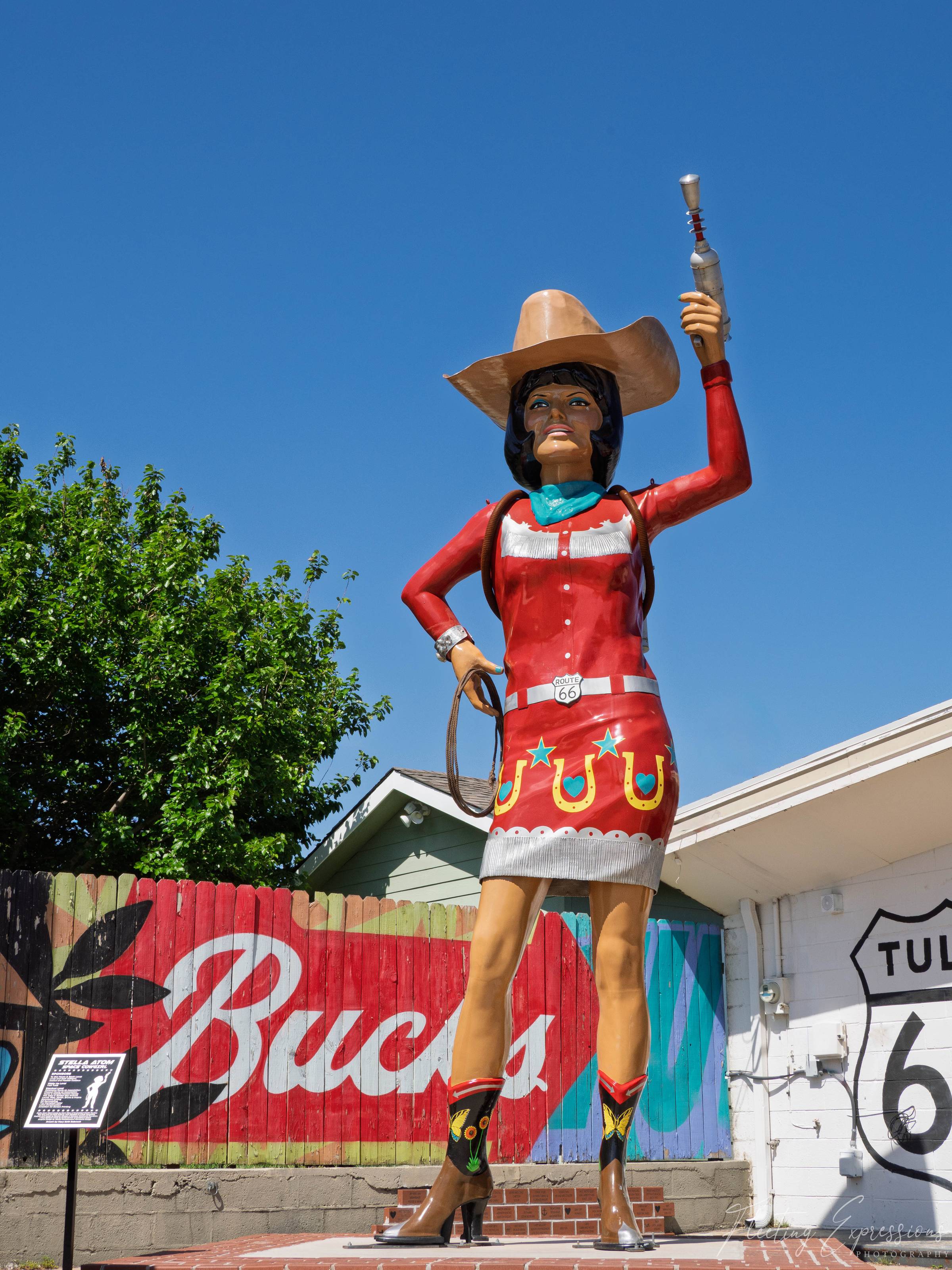 A towering cowgirl statue in a red western dress and boots raises a silver raygun overhead, standing before a fence painted with “Buck’s.” 