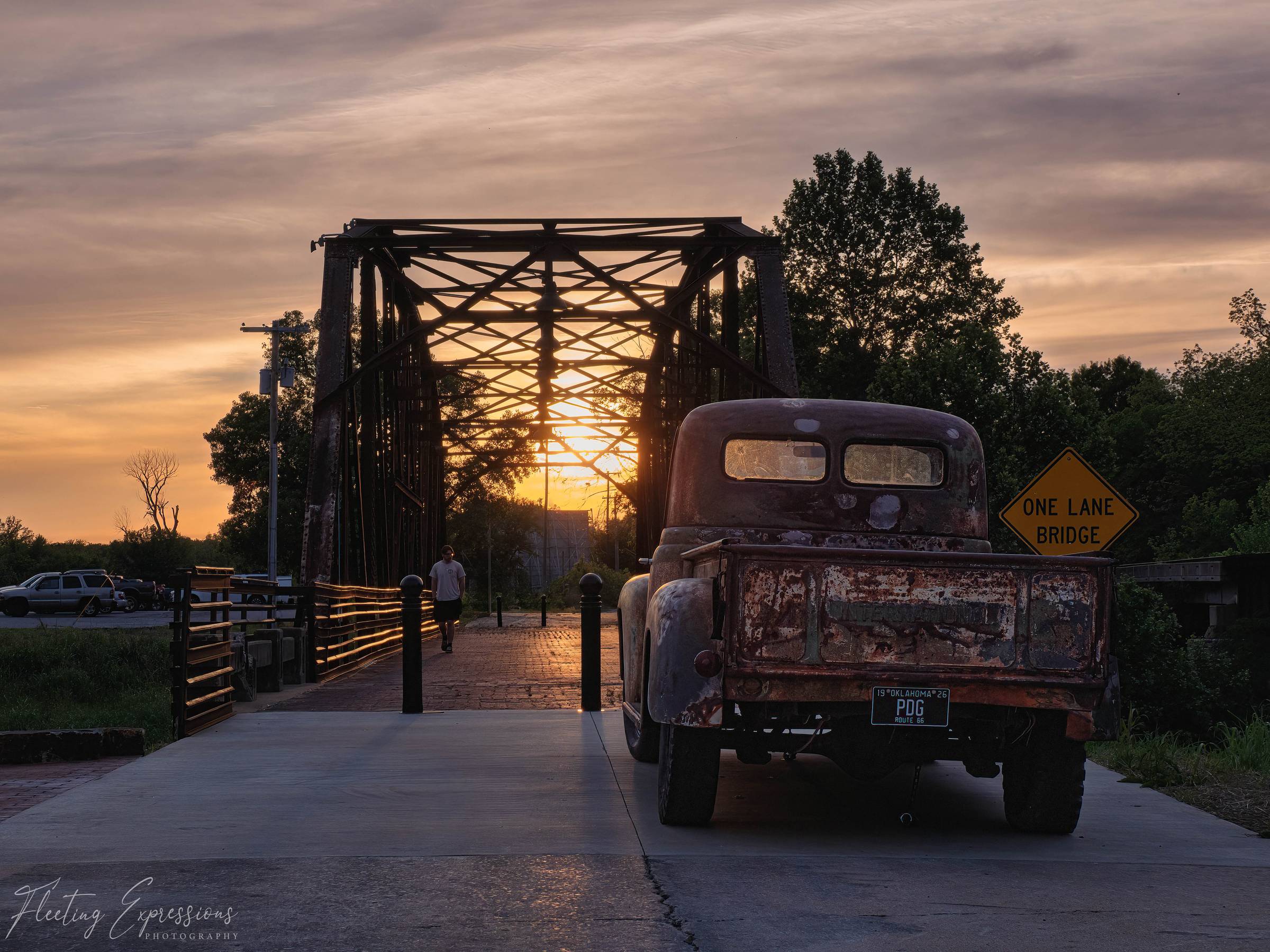 Steel truss bridge at sunset with the sun dropping through the trusses, an old rusted truck parked on the brick deck, and a One Lane Bridge sign to the right.