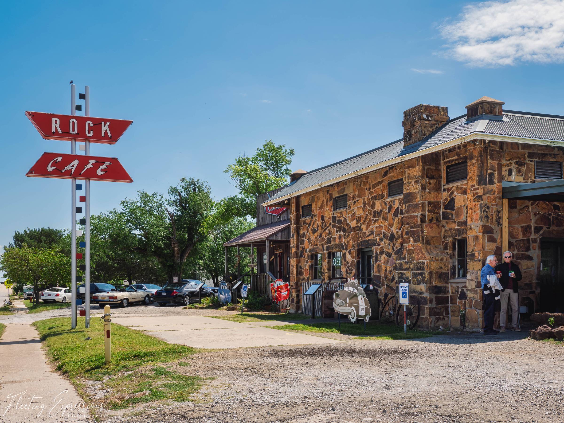 The iconic red arrow signs for the Rock Cafe in Stroud, Oklahoma, point toward the rustic sandstone building with two people standing outside under a bright blue sky.
