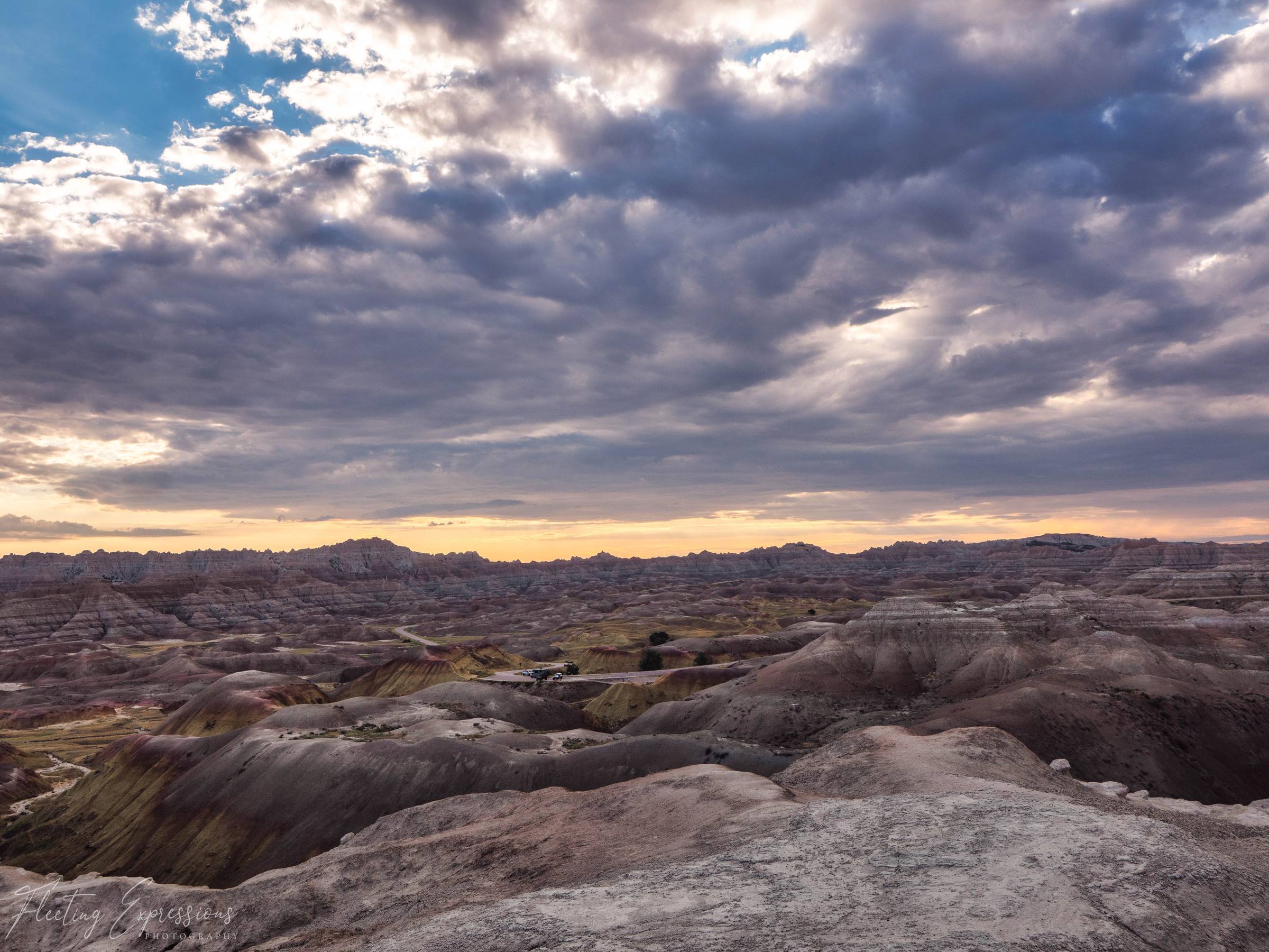 Badlands valley blanketed in heavy purple and gray storm clouds, with a paved road and a few parked vehicles visible below.