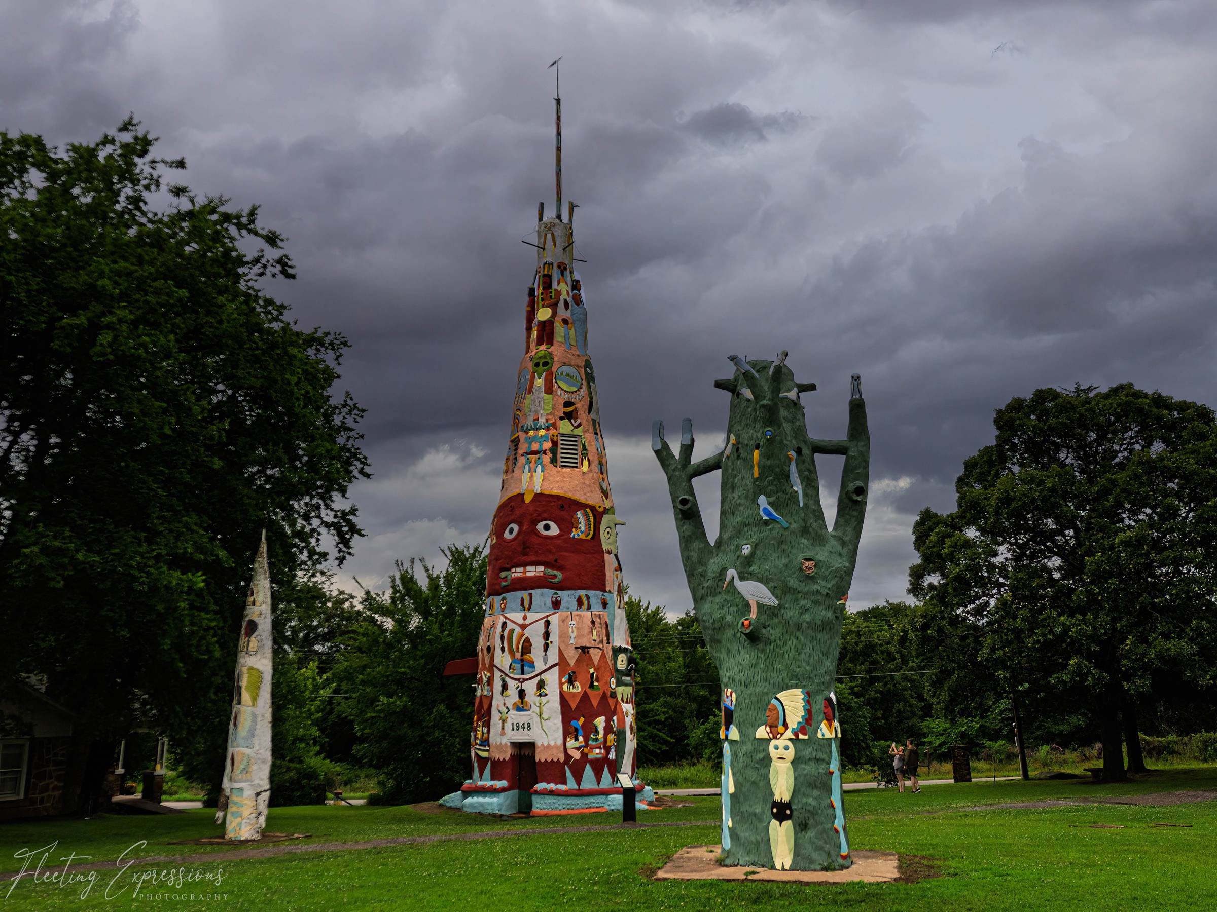 A tall painted concrete totem pole and a green carved tree sculpture stand on grass under heavy storm clouds.
