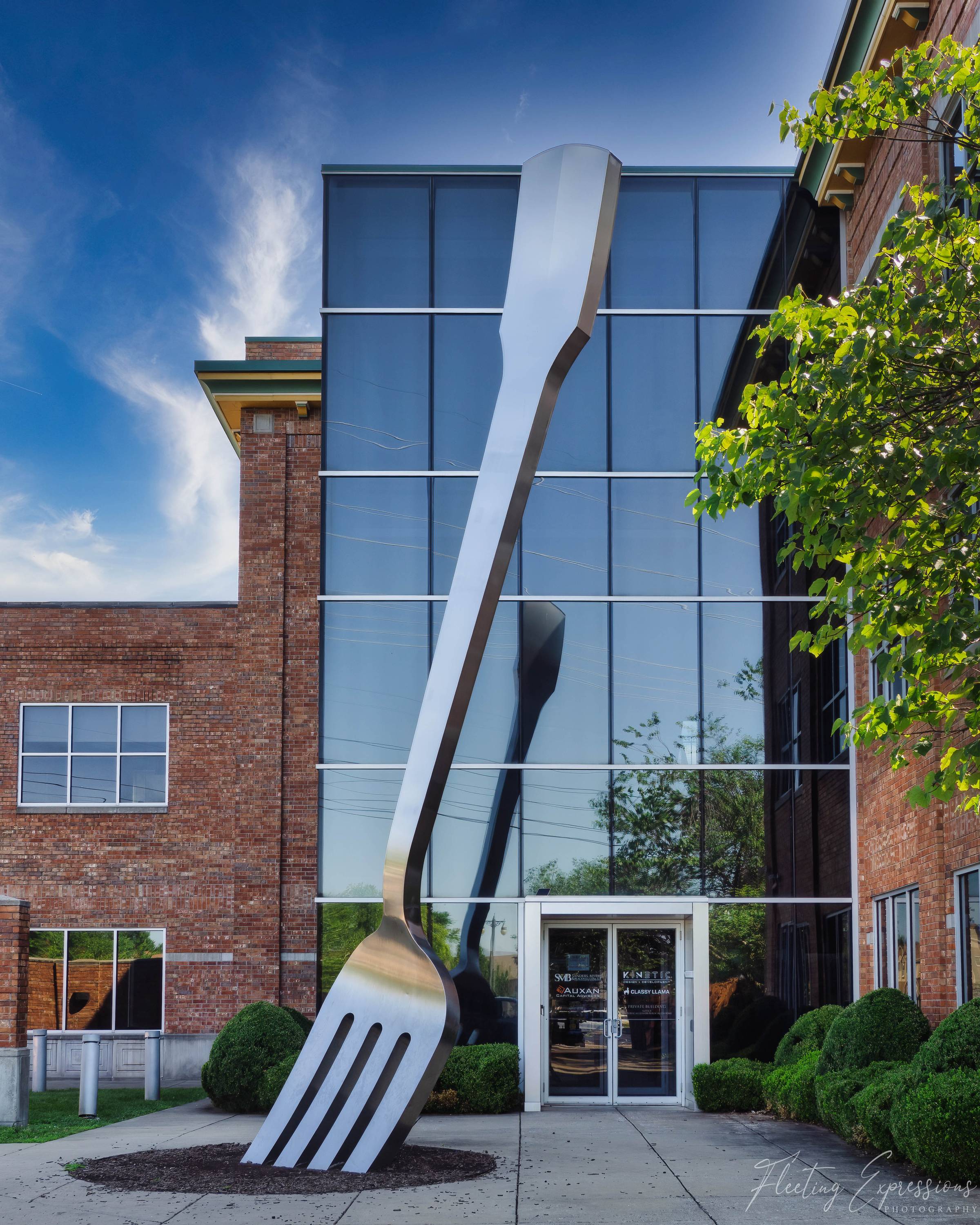 A 35-foot stainless steel fork stands upright on a sidewalk in front of a three-story glass and brick office building, dwarfing the entrance doors and reflecting sunlight along its handle.