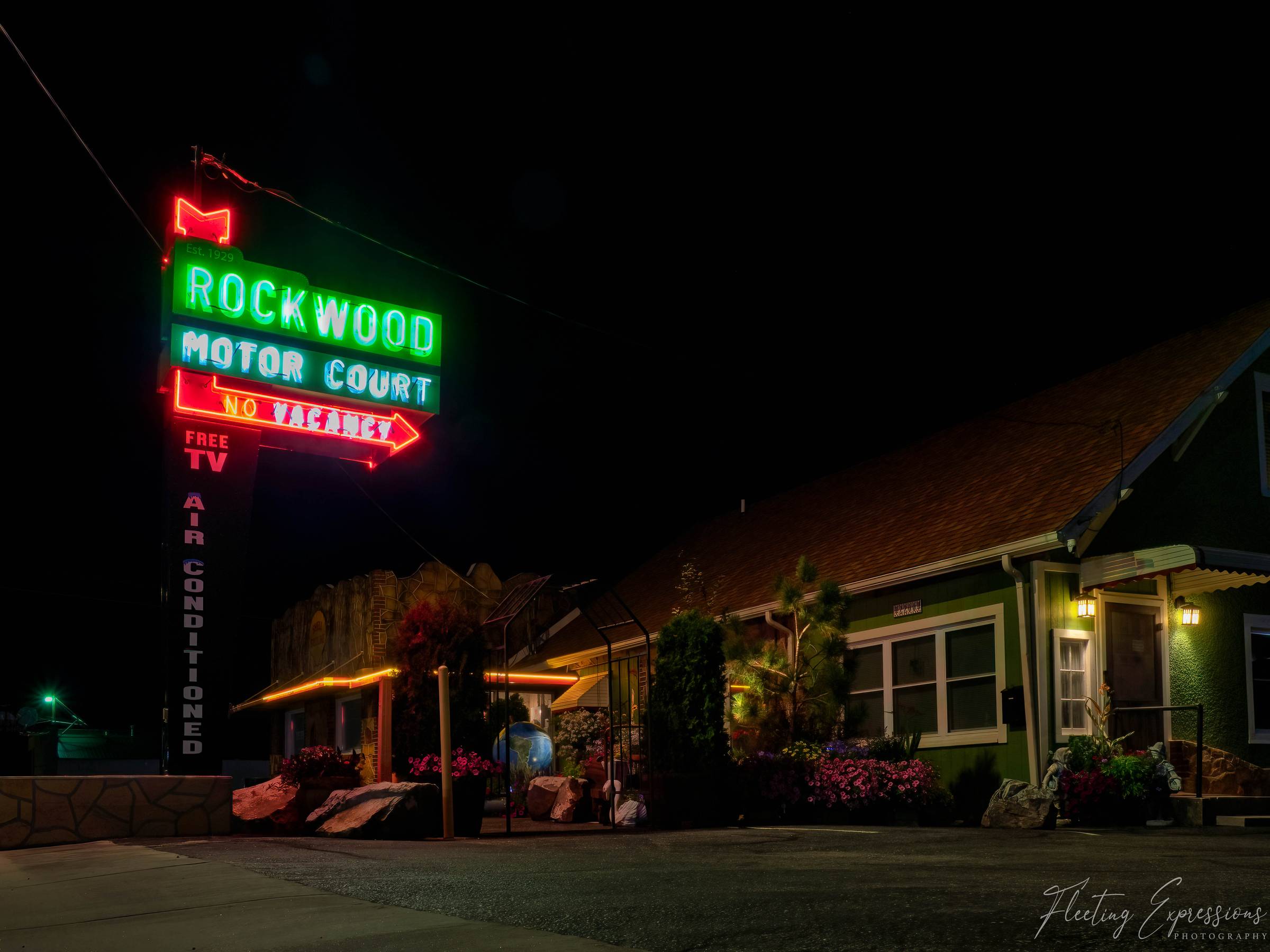 A neon sign reading "Rockwood Motor Court Est. 1929" glows green and red against a black sky above a stone motor court cottage with lit porch lamps and flower planters in front.