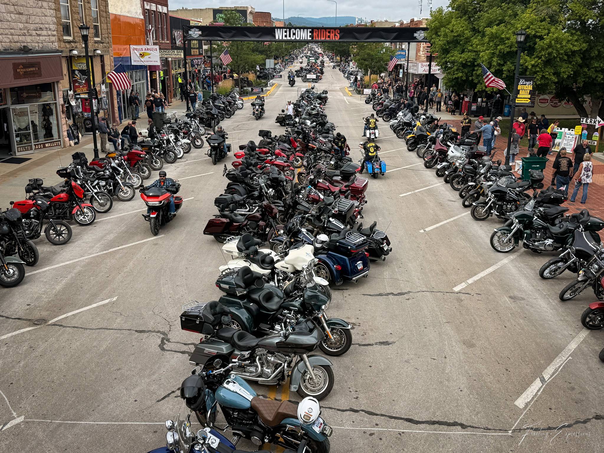 Aerial view of downtown Sturgis's Main Street packed with parked motorcycles under a Welcome Riders banner, with vendors, tourists, and storefronts lining both sides.