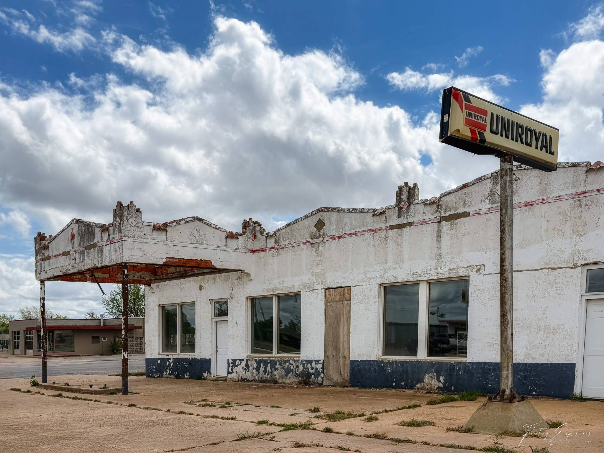A crumbling white stucco service station with a rusted metal canopy sits empty on a Route 66 roadside. A Uniroyal tire brand sign on a tall pole rises above the building against a dramatic blue sky with large white clouds. Paint peels from the lower half of the facade.