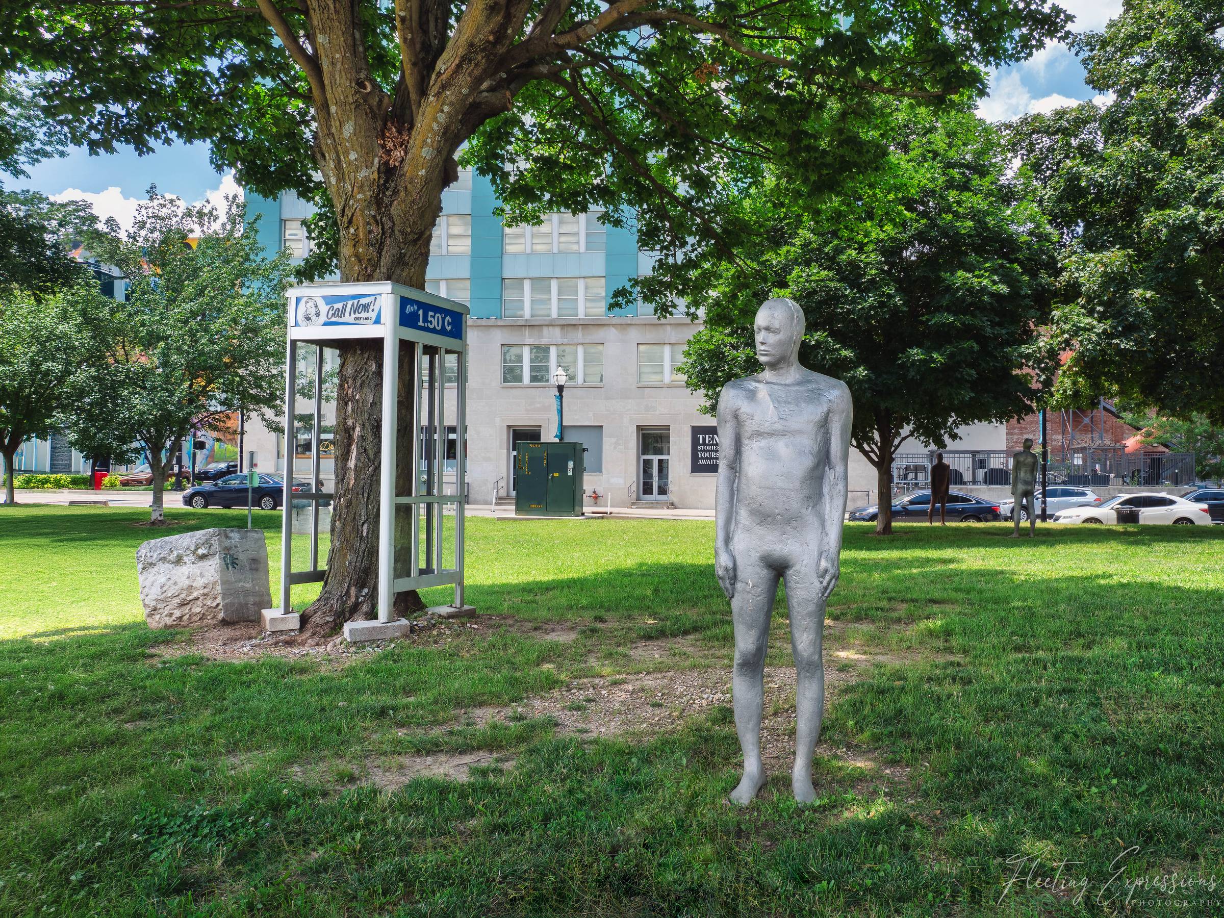 A silver human figure stands on a grassy downtown park with a vintage phone booth advertising "Call Now Only 1.50¢" behind it, with a tree growing up through the booth and additional figures visible in the distance.