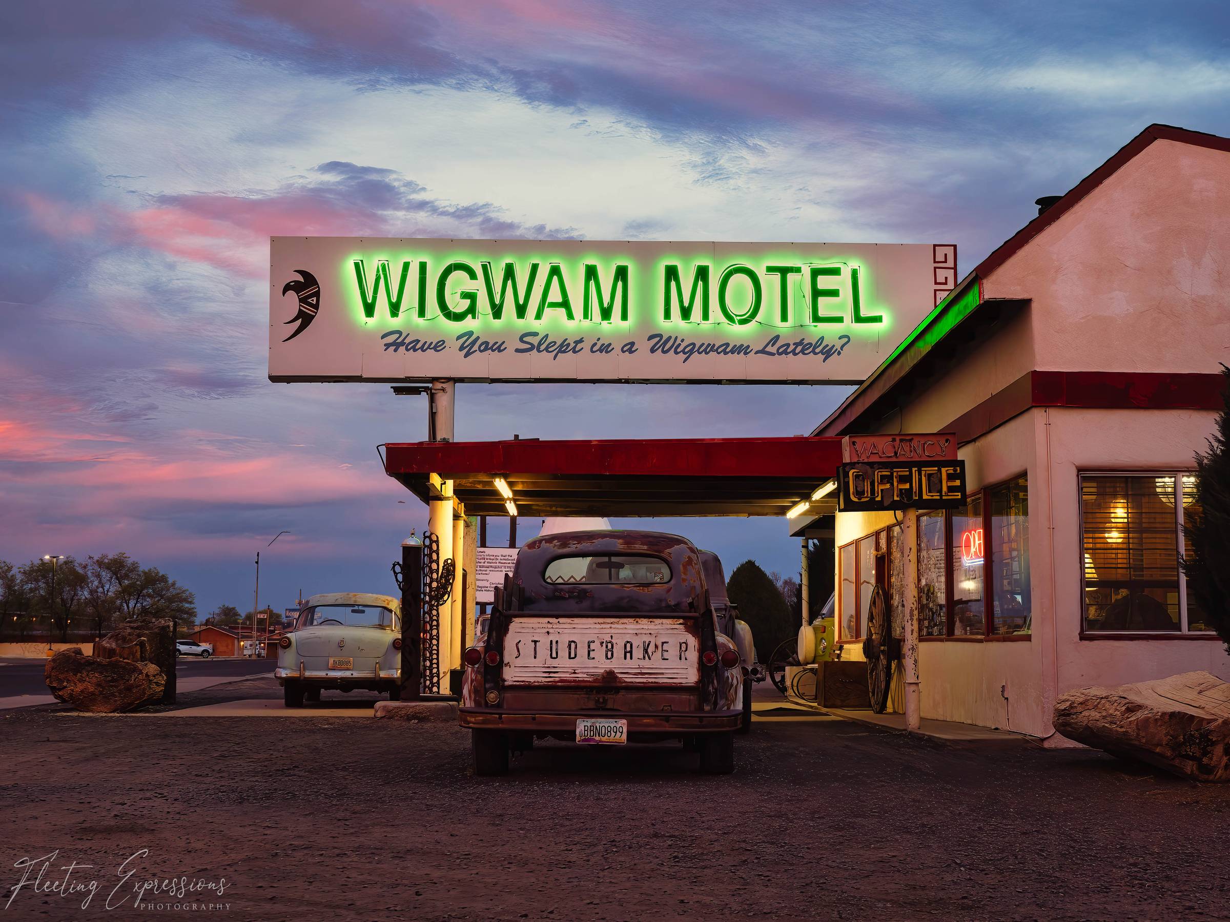 The Wigwam Motel's green neon sign glows at dusk in Holbrook, Arizona, with a rusted Studebaker parked beneath the canopy and the office light burning inside.