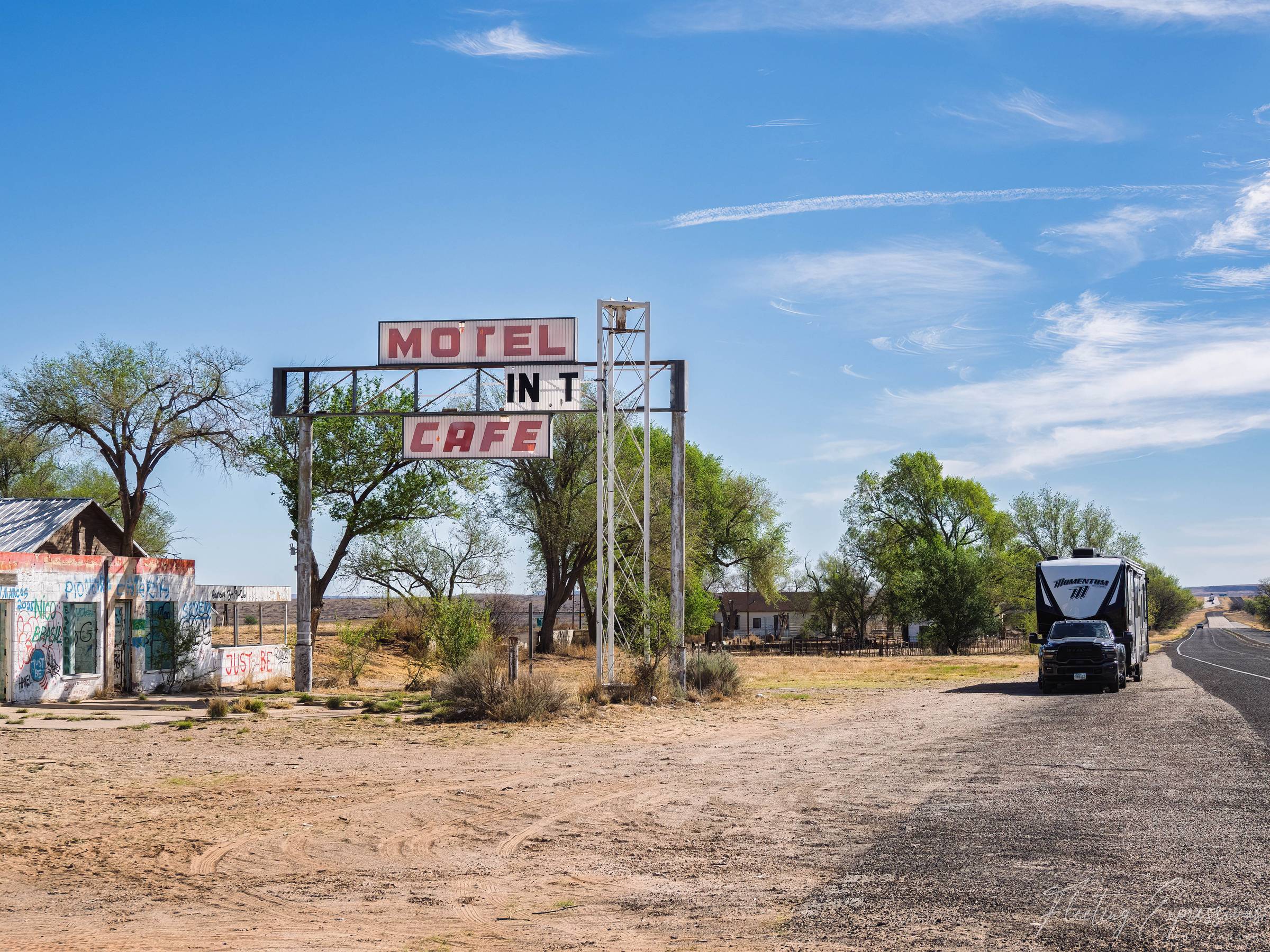 A weathered "Motel" and "Cafe" sign on a tall metal frame in the Glenrio ghost town, with a graffitied building on the left reading "Just Be" and a black truck towing a white RV parked on the right.