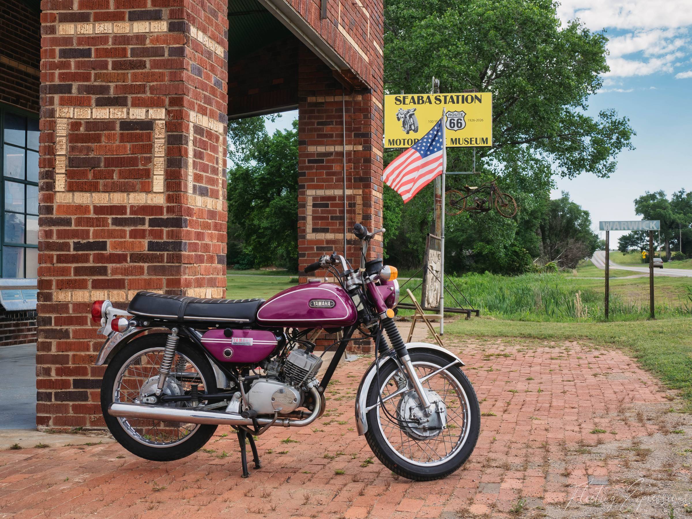 A vintage purple Yamaha motorcycle parked on brick pavers in front of the historic Seaba Station building in Warwick, Oklahoma, with the Route 66 Centennial sign and an American flag visible in the background.
