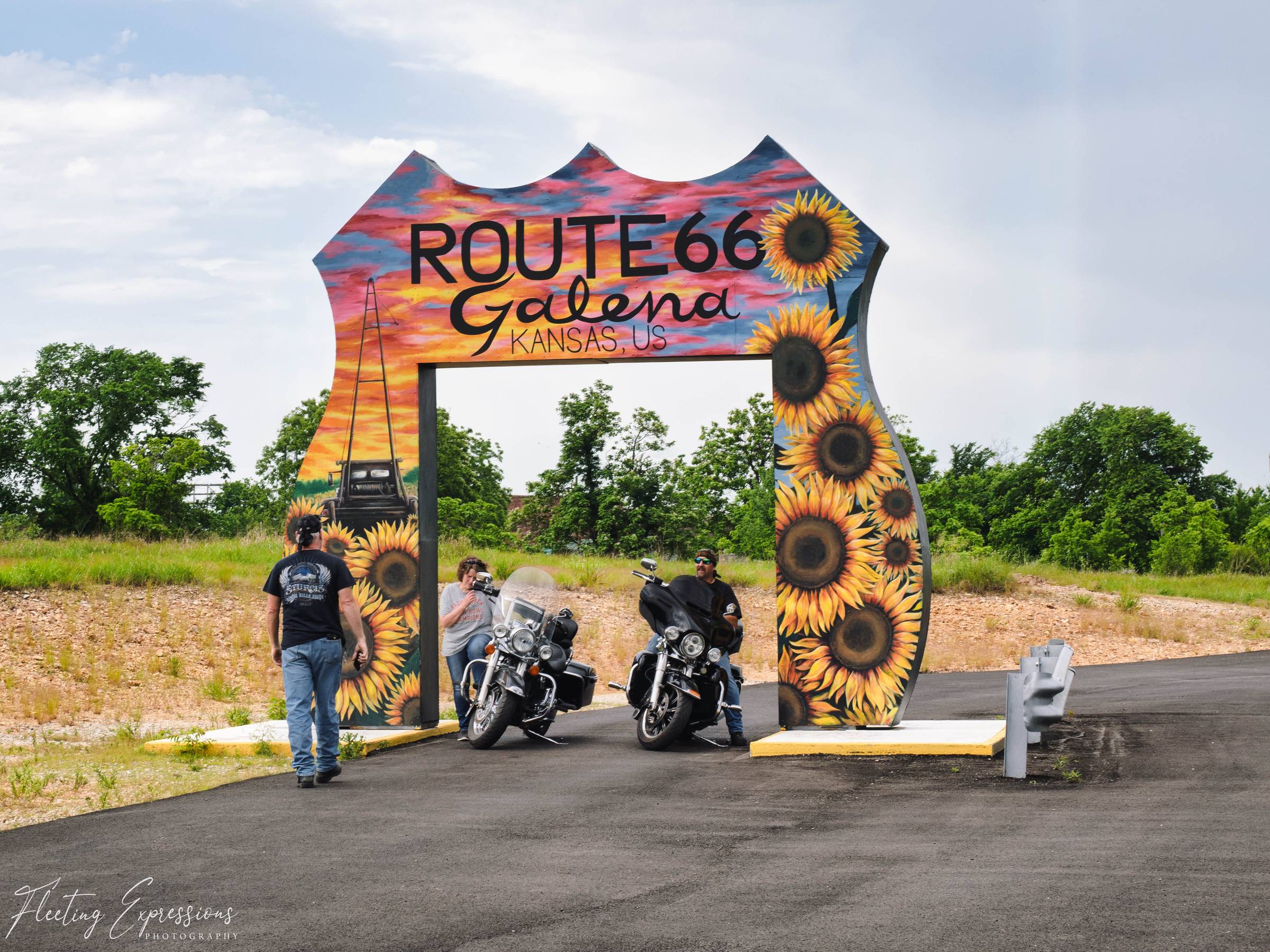 Two motorcycles park beneath a tall painted arch reading "Route 66 Galena Kansas, US," decorated with sunflowers and a sunset.