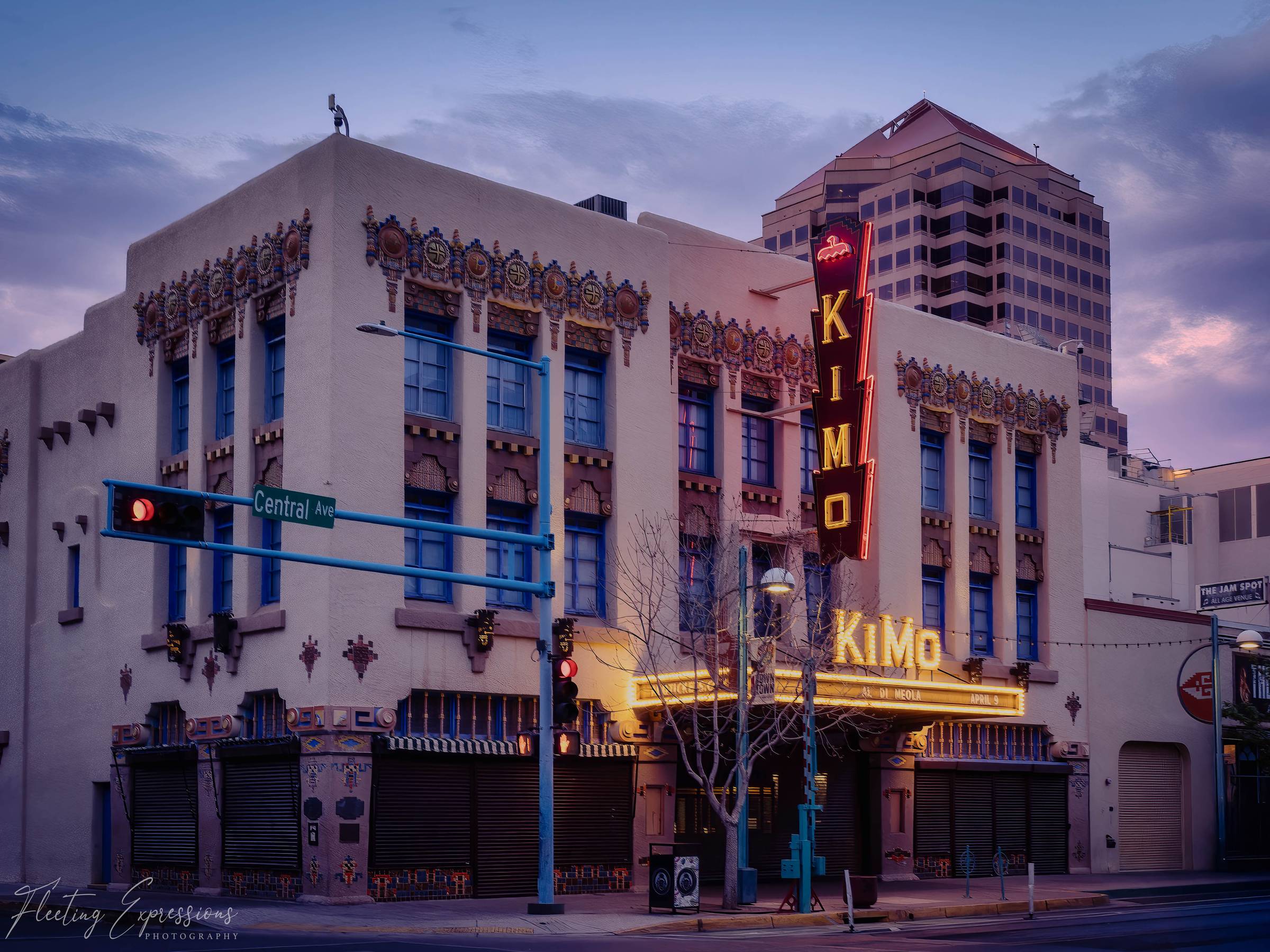 The KiMo Theatre on Central Avenue in Albuquerque, New Mexico, lit up at dusk with its Pueblo Deco facade and neon vertical sign glowing against a purple sky.