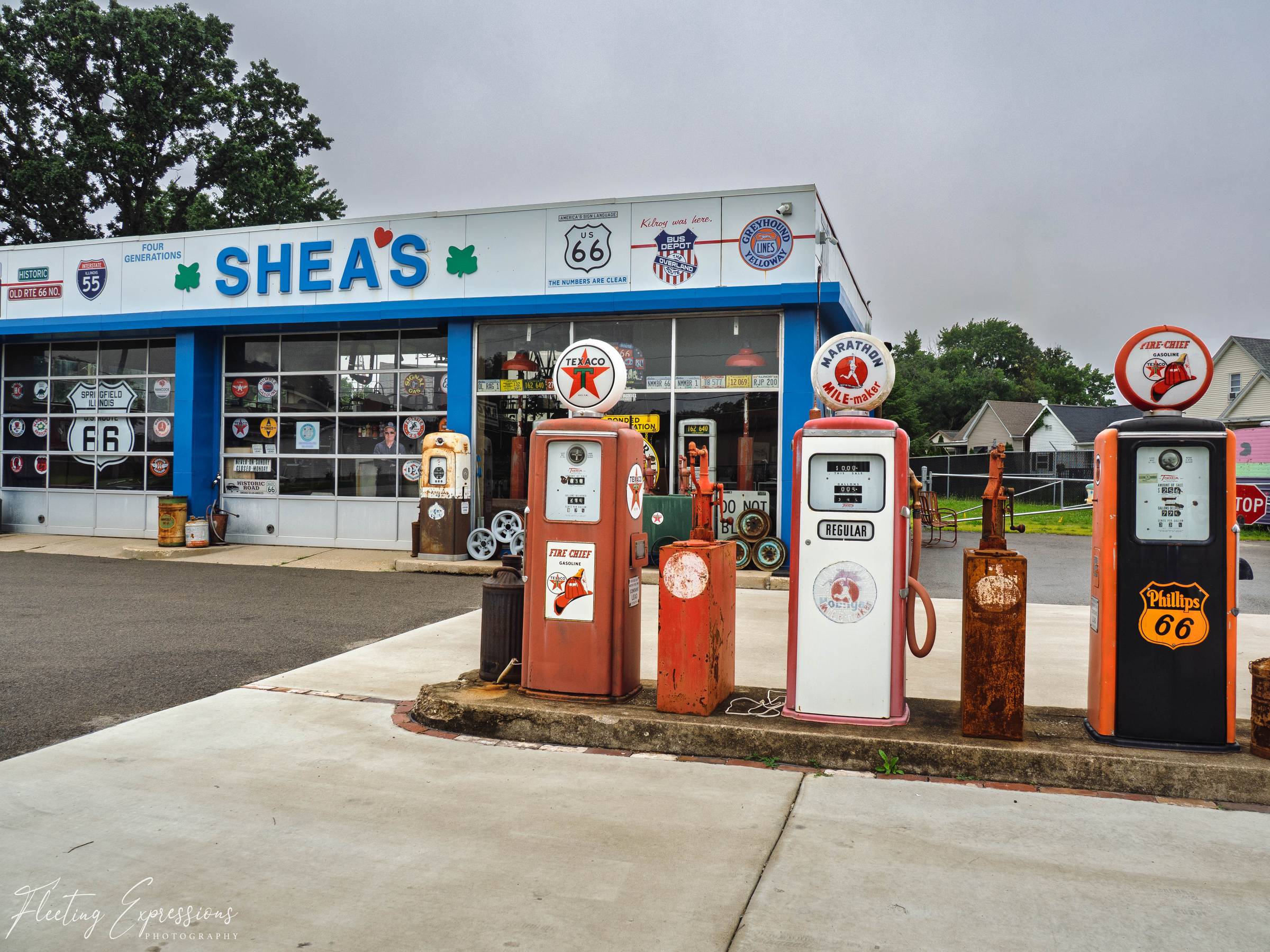 Blue-trimmed storefront covered in vintage Route 66 and gasoline signs; four antique gas pumps stand in the foreground, including red Fire Chief Texaco, white Marathon, and orange Phillips 66.