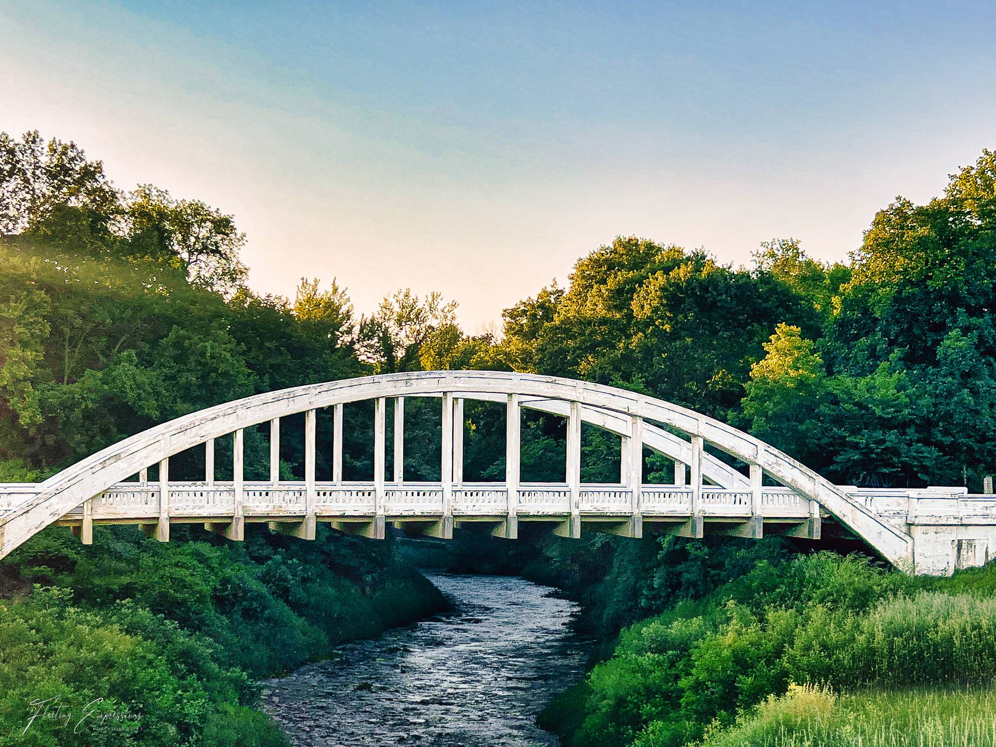A white concrete arch bridge spans a rocky creek lined with green brush, its single span lit by low evening sun.