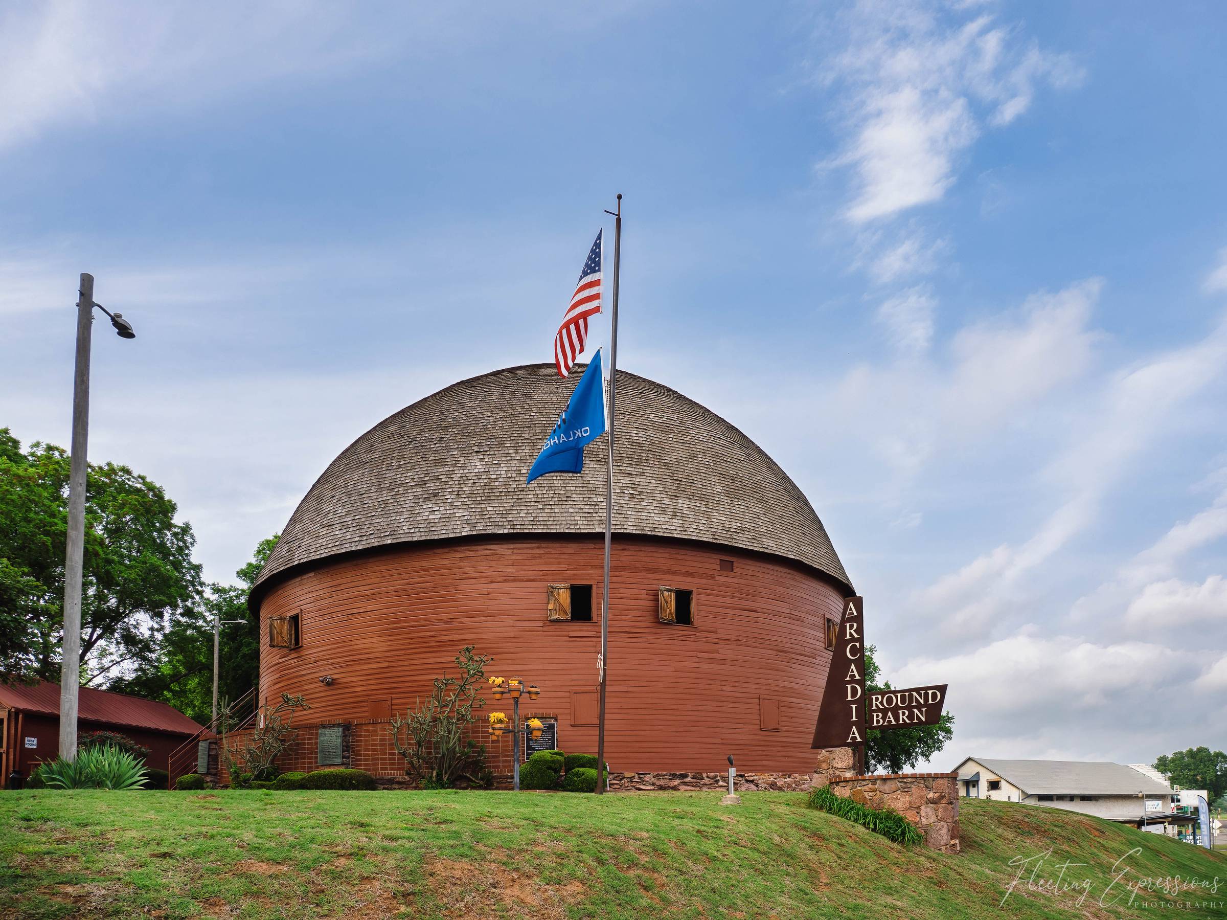  The Arcadia Round Barn, a circular red wooden structure with a domed roof, flying an American flag and Oklahoma state flag, surrounded by green grass in Arcadia, Oklahoma.