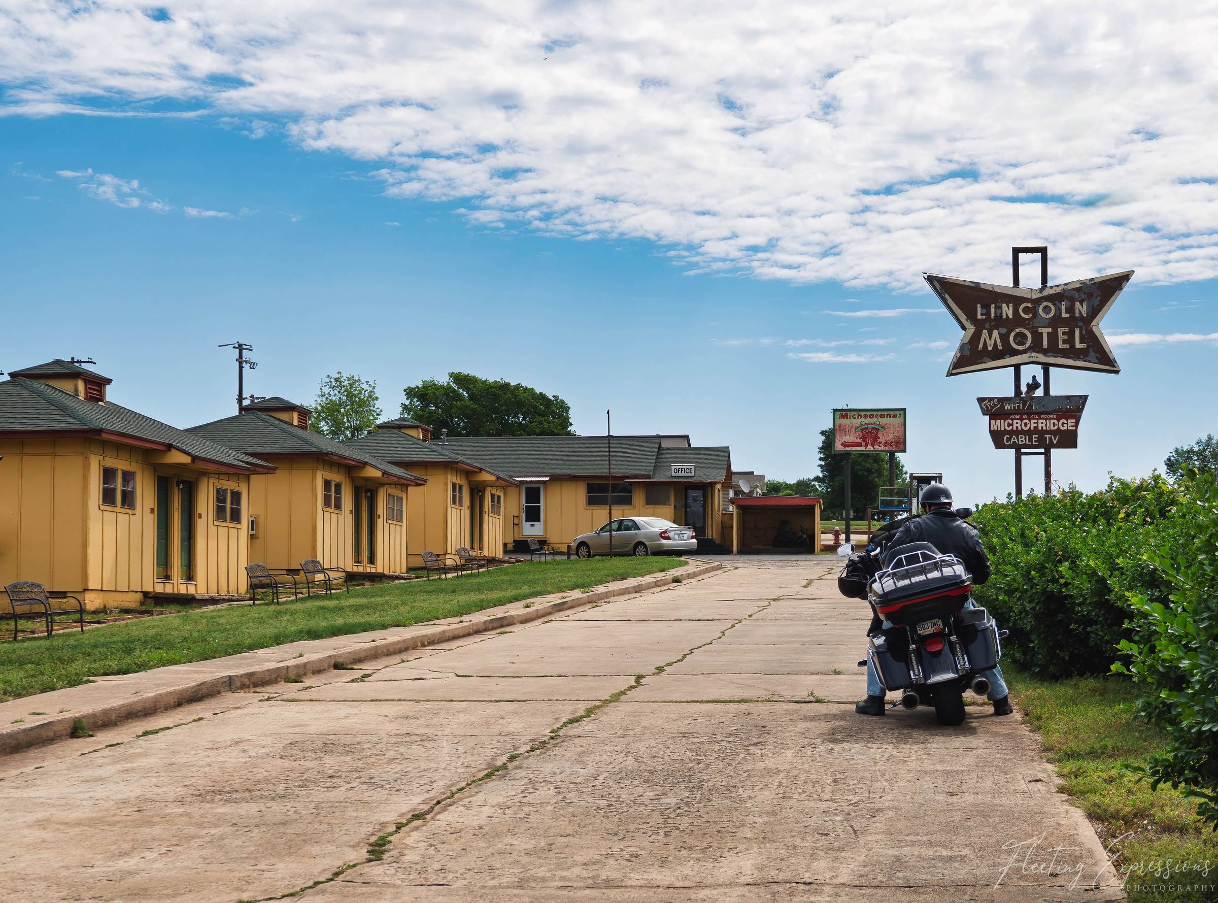 A motorcyclist approaches the weathered neon sign for the Lincoln Motel in Chandler, Oklahoma, with a row of classic yellow cottage-style motel rooms lining the driveway under a partly cloudy sky.