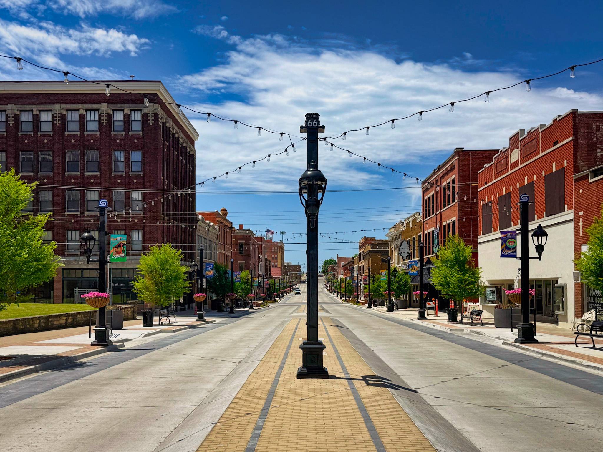 Empty brick main street in historic downtown Sapulpa, lined with old brick buildings and string lights, a Route 66 sign on a lamppost in the center, under a bright blue sky.