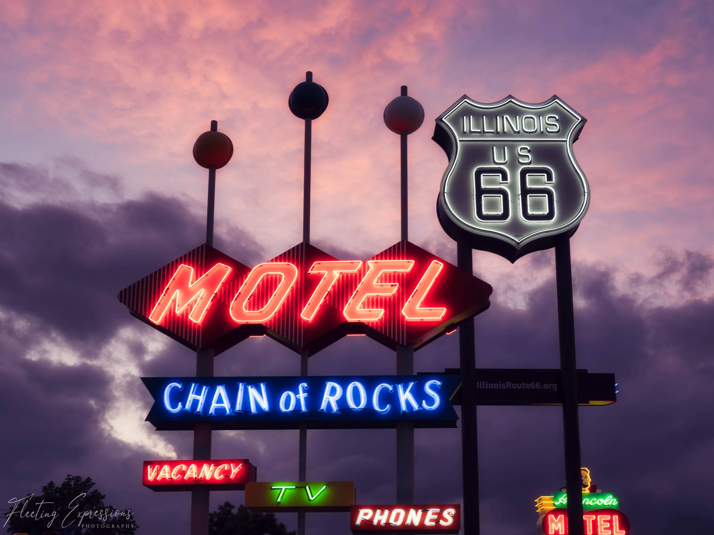 Neon signs reading Motel, Chain of Rocks, and Illinois US 66 glow against a pink dusk sky.
