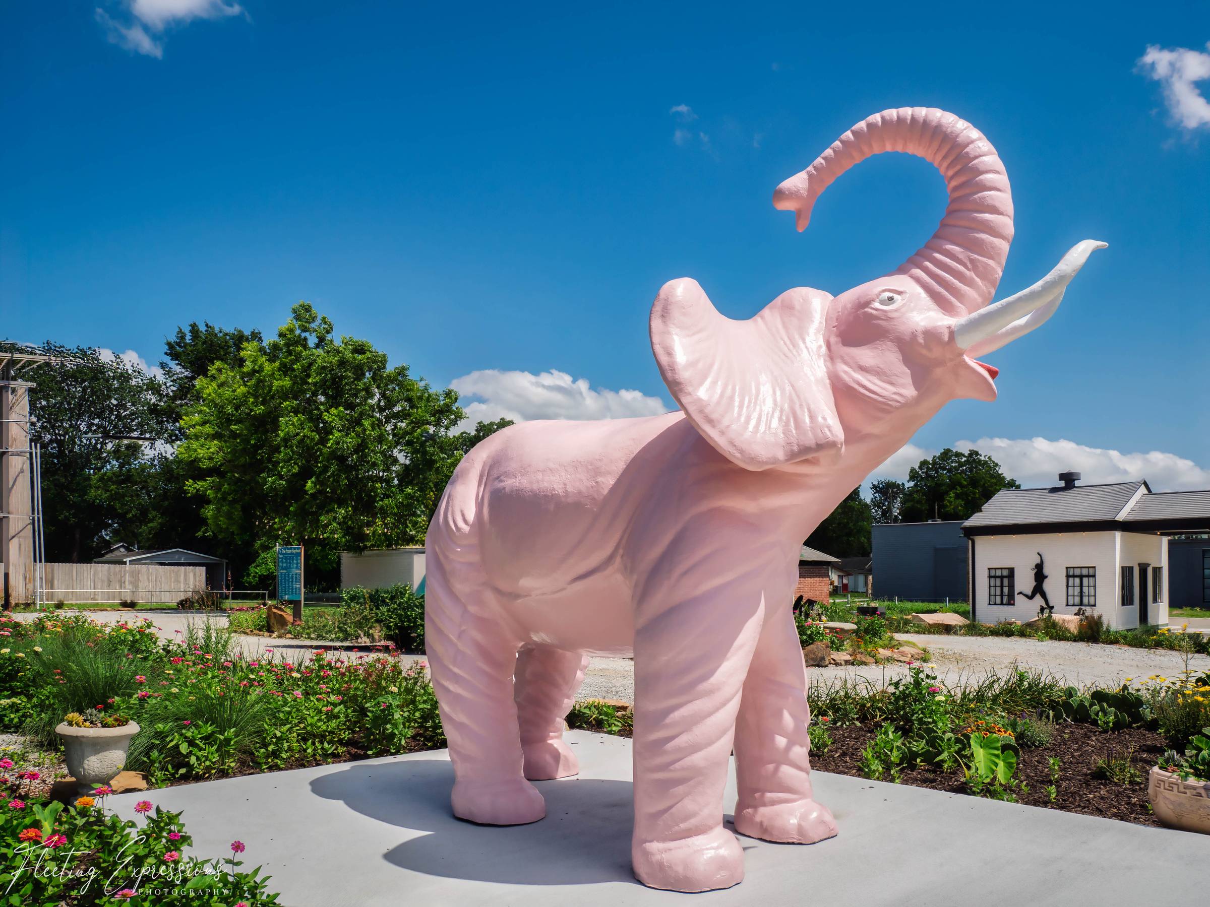 A large pink elephant statue raises its trunk against a blue sky, surrounded by flowers near a restored gas station.