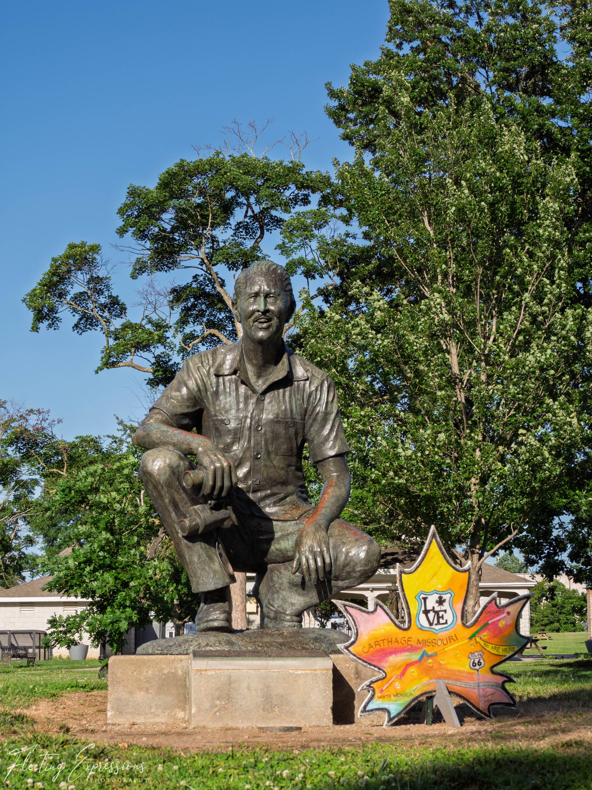 Bronze statue of Marlin Perkins in a Carthage park beside a painted Route 66 maple leaf sign