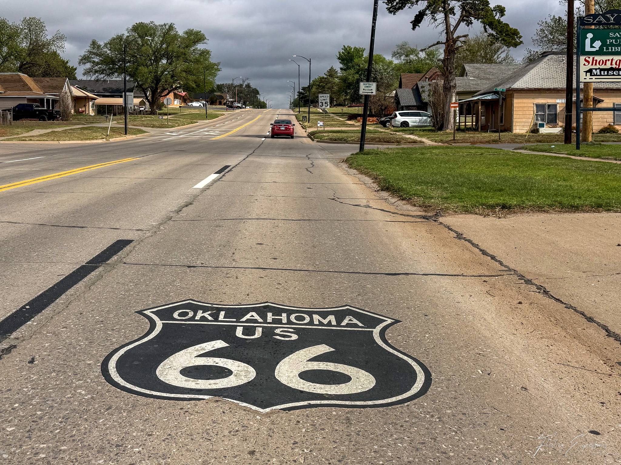 An Oklahoma Route 66 shield is painted in black and white directly on the pavement of a two-lane road in Sayre, Oklahoma. The road stretches ahead under a heavy gray storm sky, with small town buildings and trees lining both sides. A lone red car is visible in the distance.