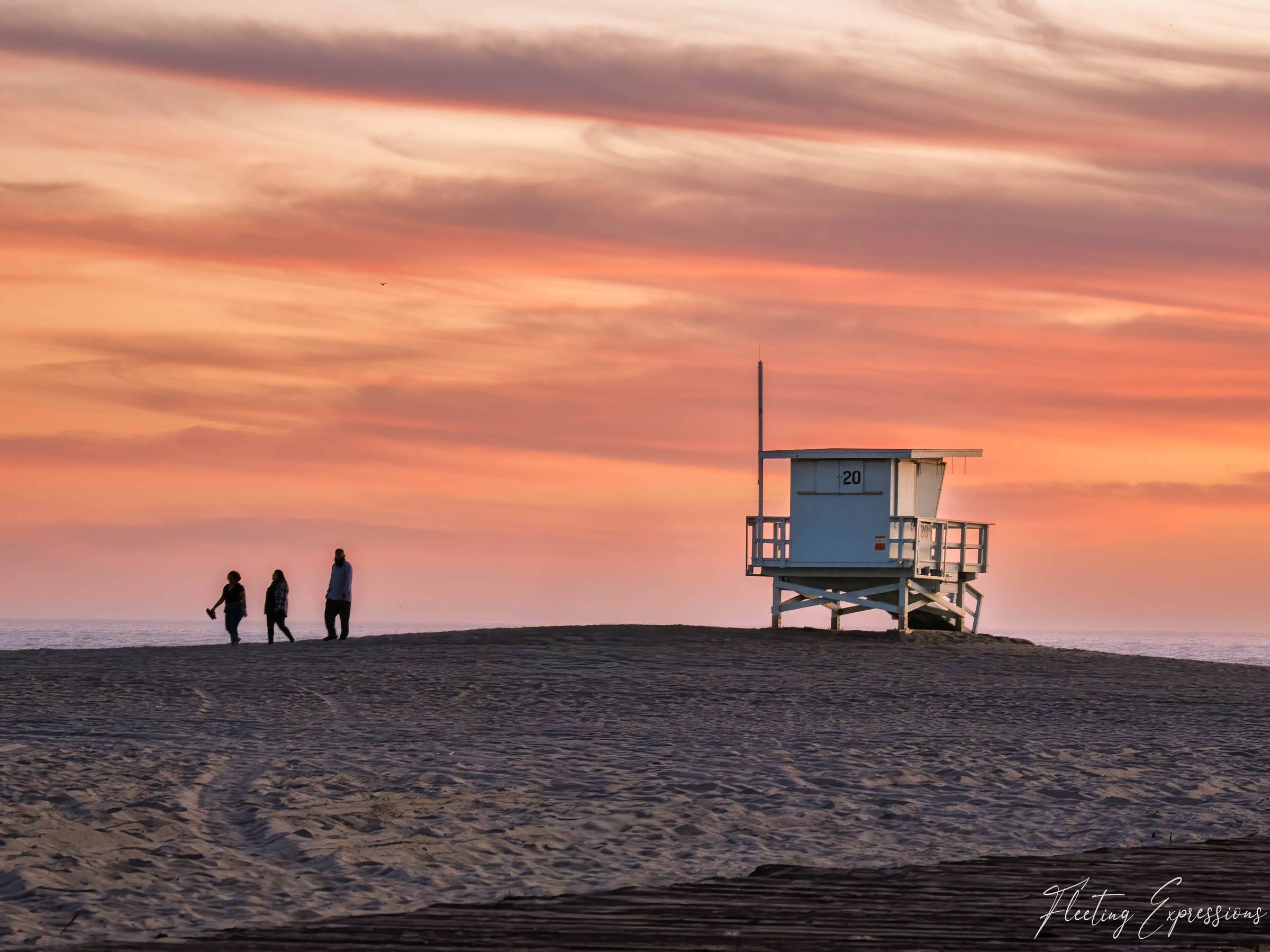 Silhouetted lifeguard tower and three people walking along a sandy ridge at sunset in Santa Monica, with pink and orange clouds over the Pacific Ocean.