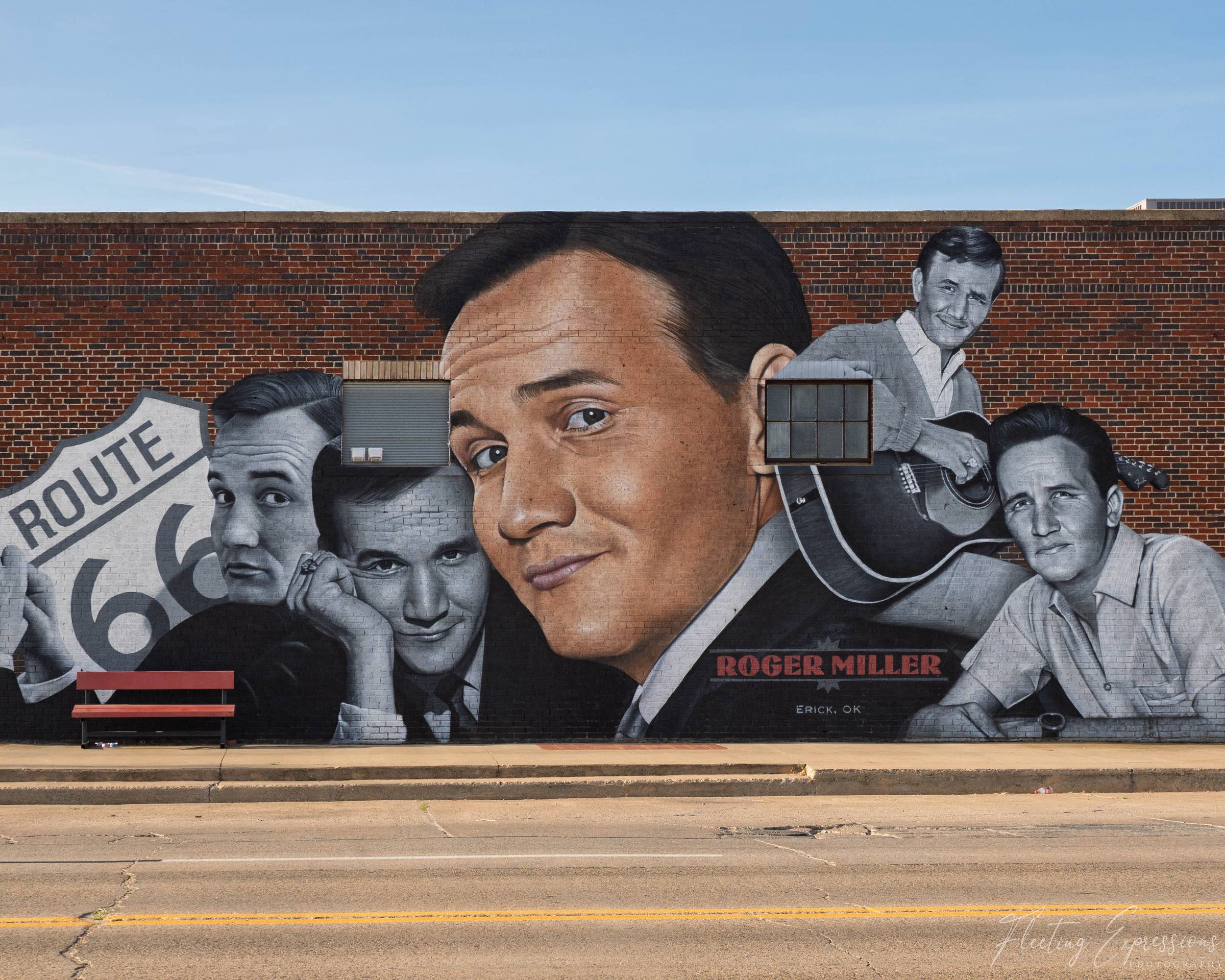 A large photorealistic mural covers a full brick wall in Erick, Oklahoma, depicting country music legend Roger Miller at various stages of his career in black and white, with a central color portrait. A Route 66 shield appears on the left side of the mural. A red bench sits on the sidewalk below.