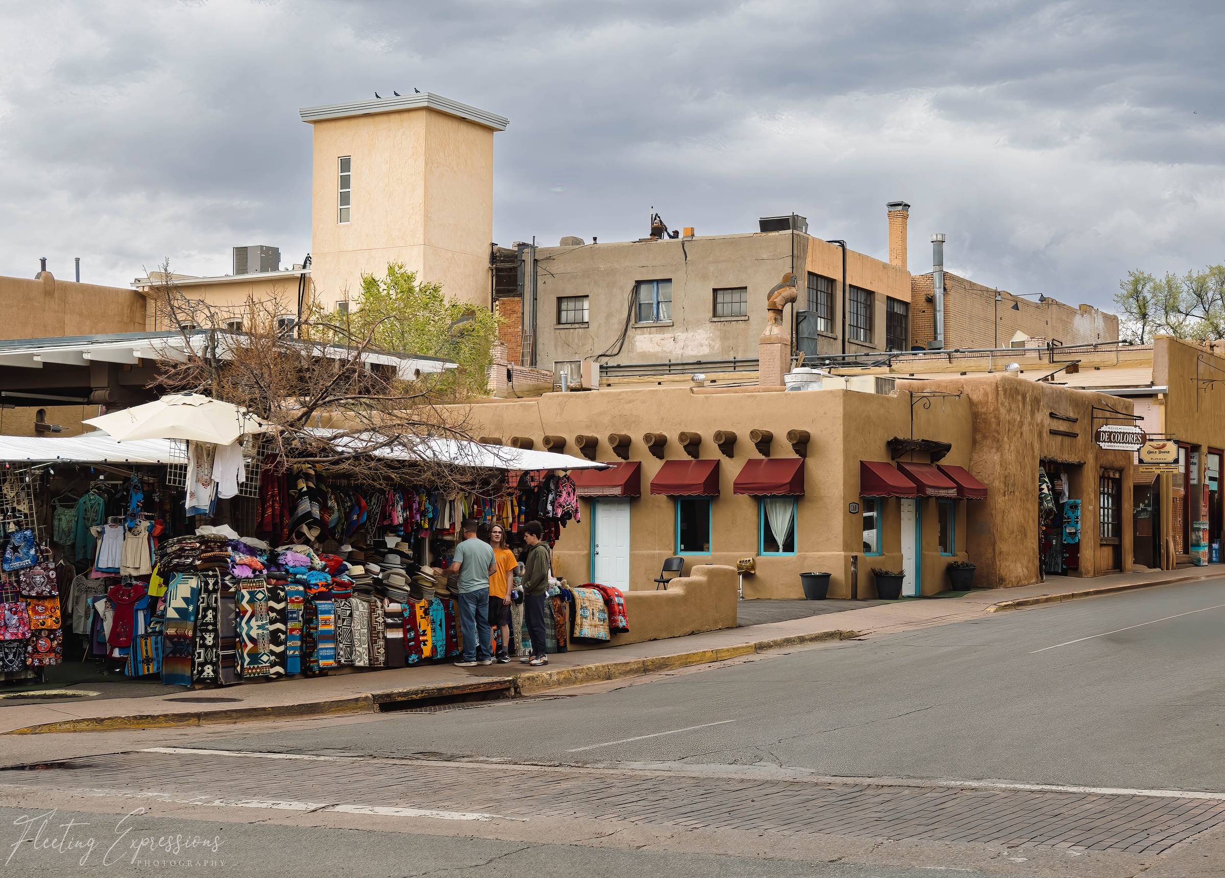 A colorful outdoor market vendor displaying textiles on a street corner in historic downtown Santa Fe, New Mexico, with adobe architecture and a stormy sky overhead.