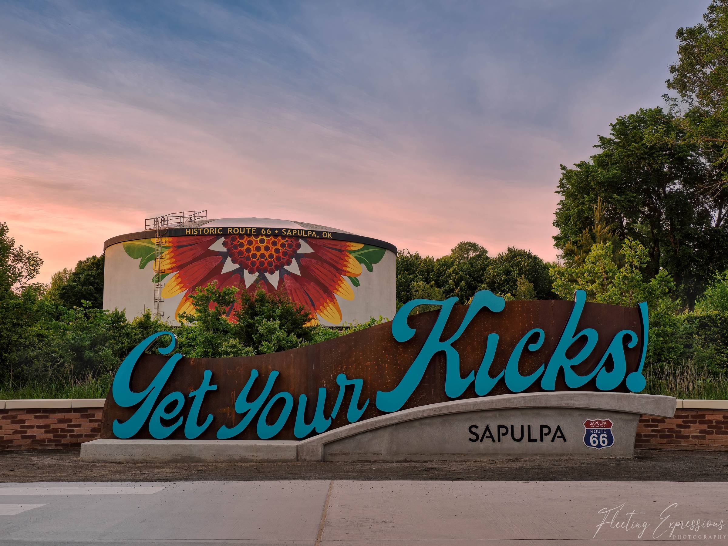  Large teal cursive "Get Your Kicks!" sign on a rusted steel base, with a water tower painted with a red and yellow Indian Blanket flower behind it, under a pink and blue sunset.