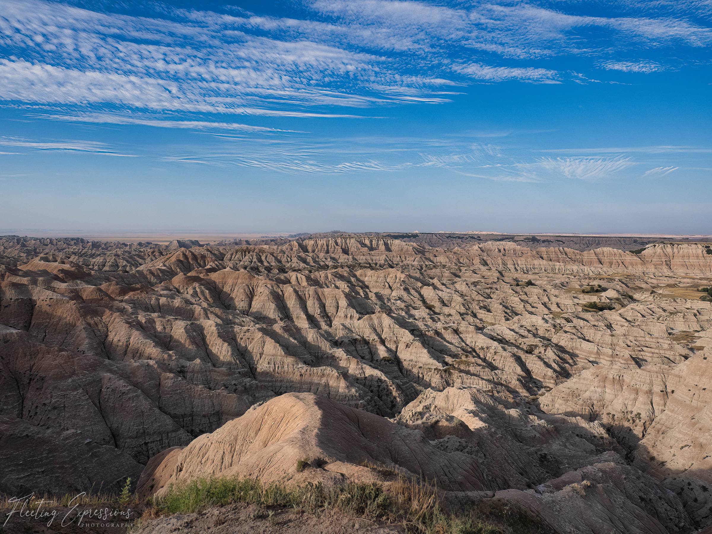 Wide view of grass capped badlands buttes stretching toward the horizon under a blue sky streaked with thin white clouds.