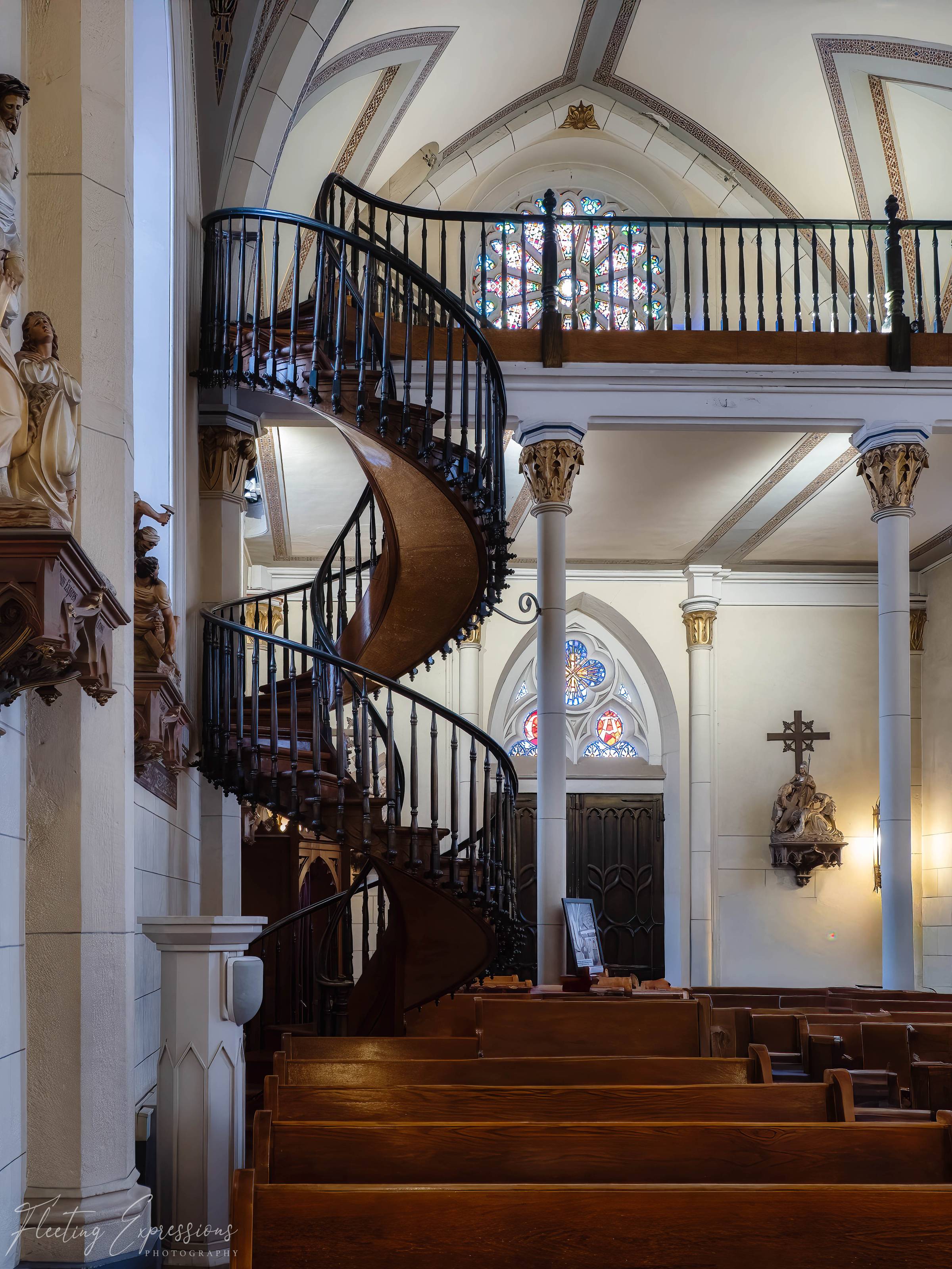 The famous spiral staircase inside Loretto Chapel in Santa Fe, New Mexico, with a rose window visible in the background.