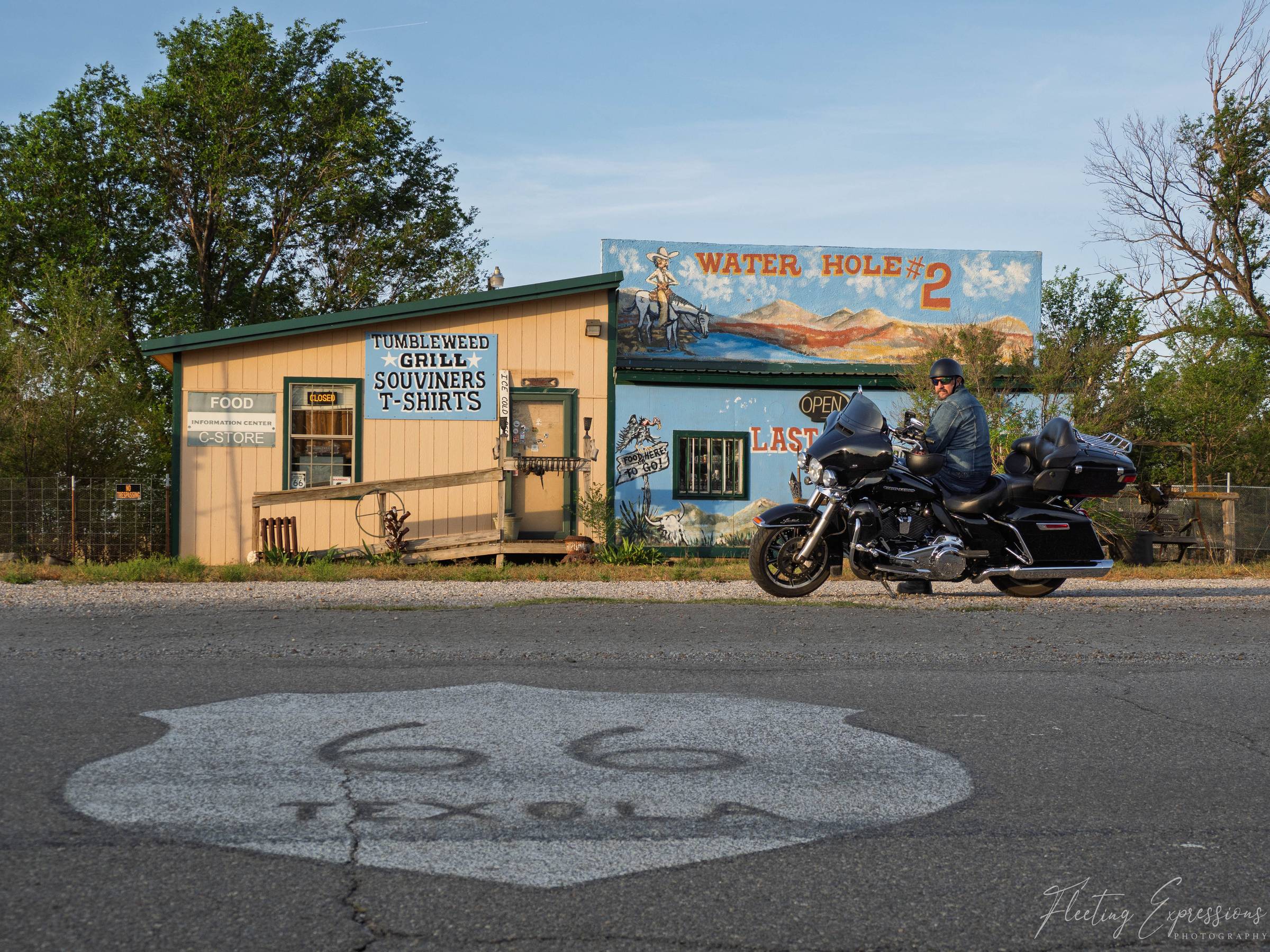 A motorcyclist in a denim jacket sits on a black Harley-Davidson in front of a faded yellow roadside building with a hand-painted mural reading "Water Hole #2" in Texola, Oklahoma. A Route 66 shield is painted on the cracked asphalt in the foreground. Ear