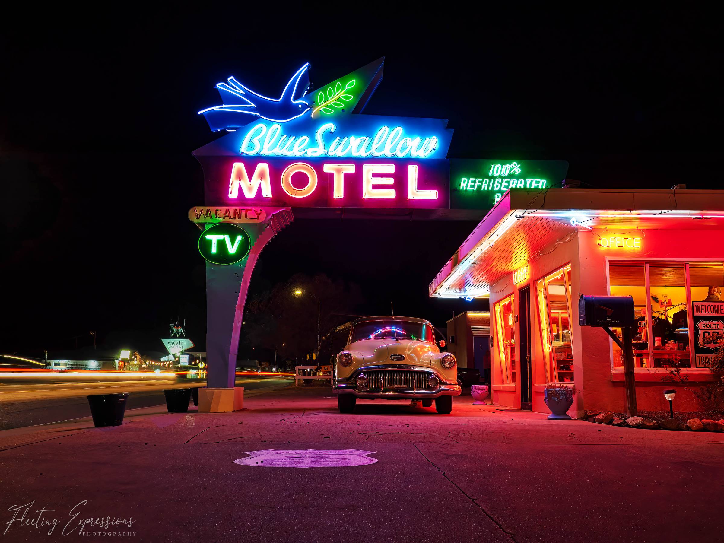 The Blue Swallow Motel neon sign glowing at night in Tucumcari, New Mexico, with a vintage car parked beneath it and light trails from passing traffic on Route 66.