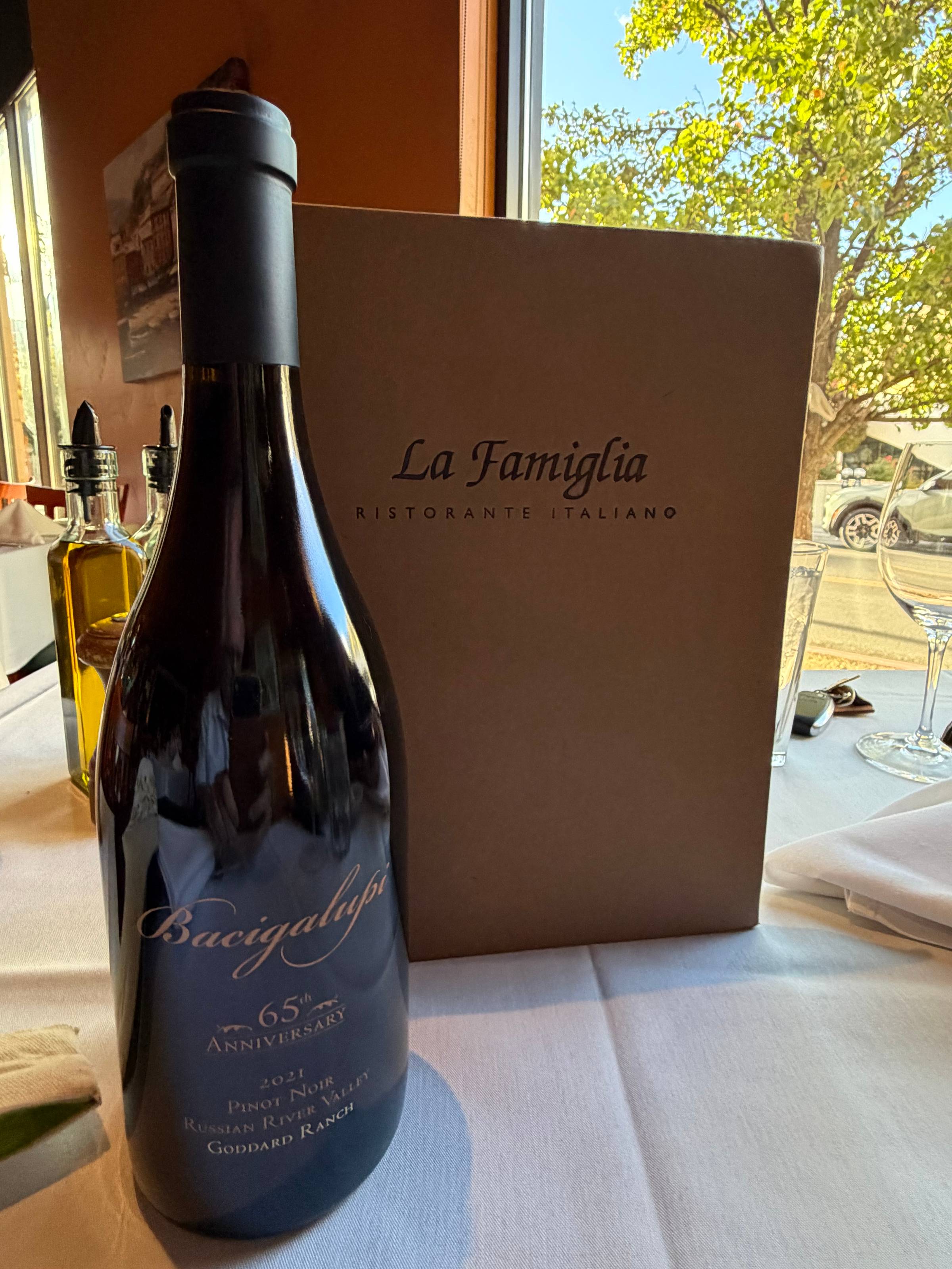 A bottle of Bacigalupi 65th Anniversary Goddard Ranch Pinot Noir standing on a white tablecloth beside a La Famiglia Ristorante Italiano menu, with olive oil cruets, a wine glass, and a sunlit window behind.
