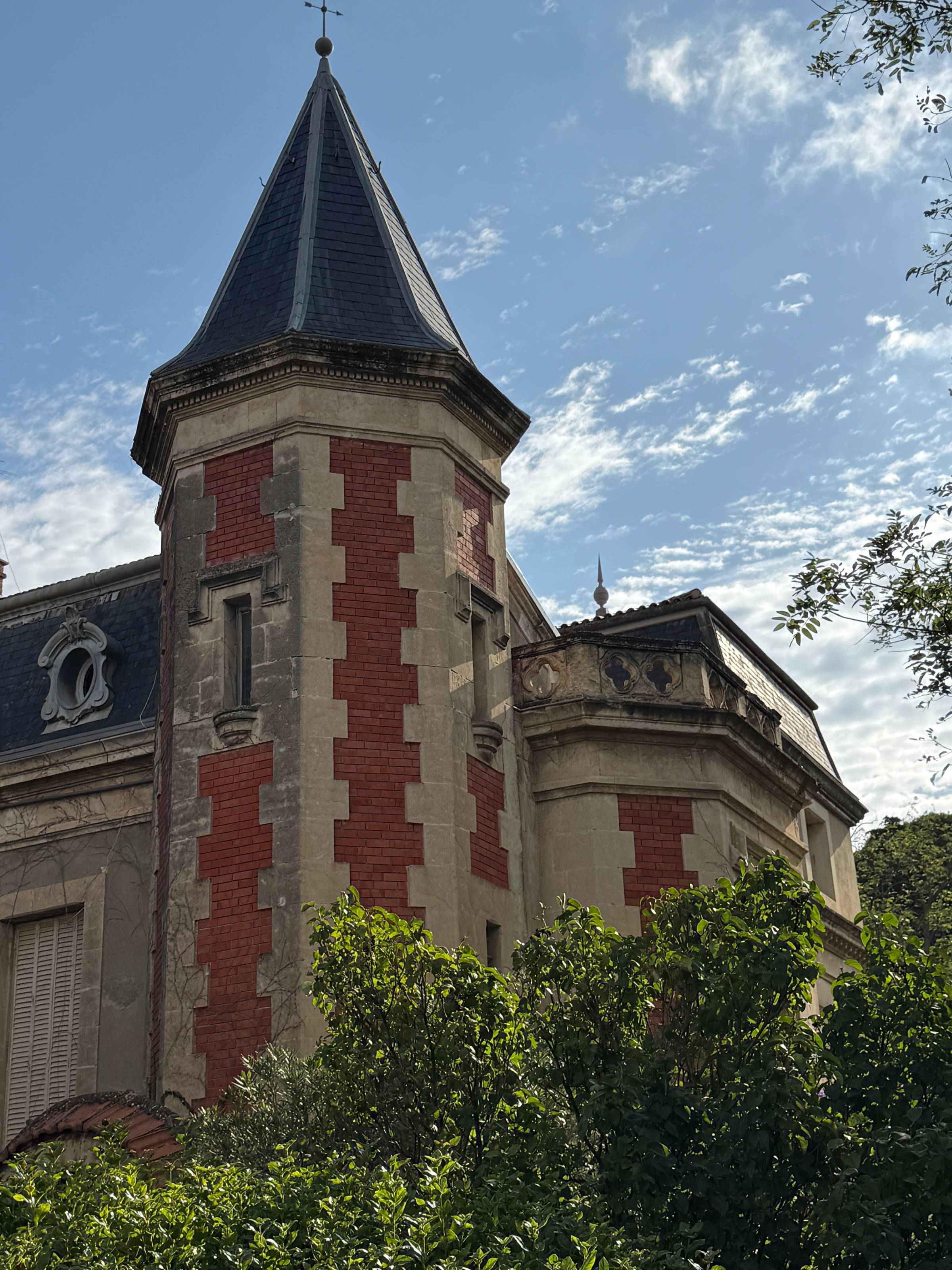 The exterior of a French wine estate in Châteauneuf-du-Pape, featuring a classic stone turret with red brick detailing and a dark slate roof, framed by green foliage under a blue sky.