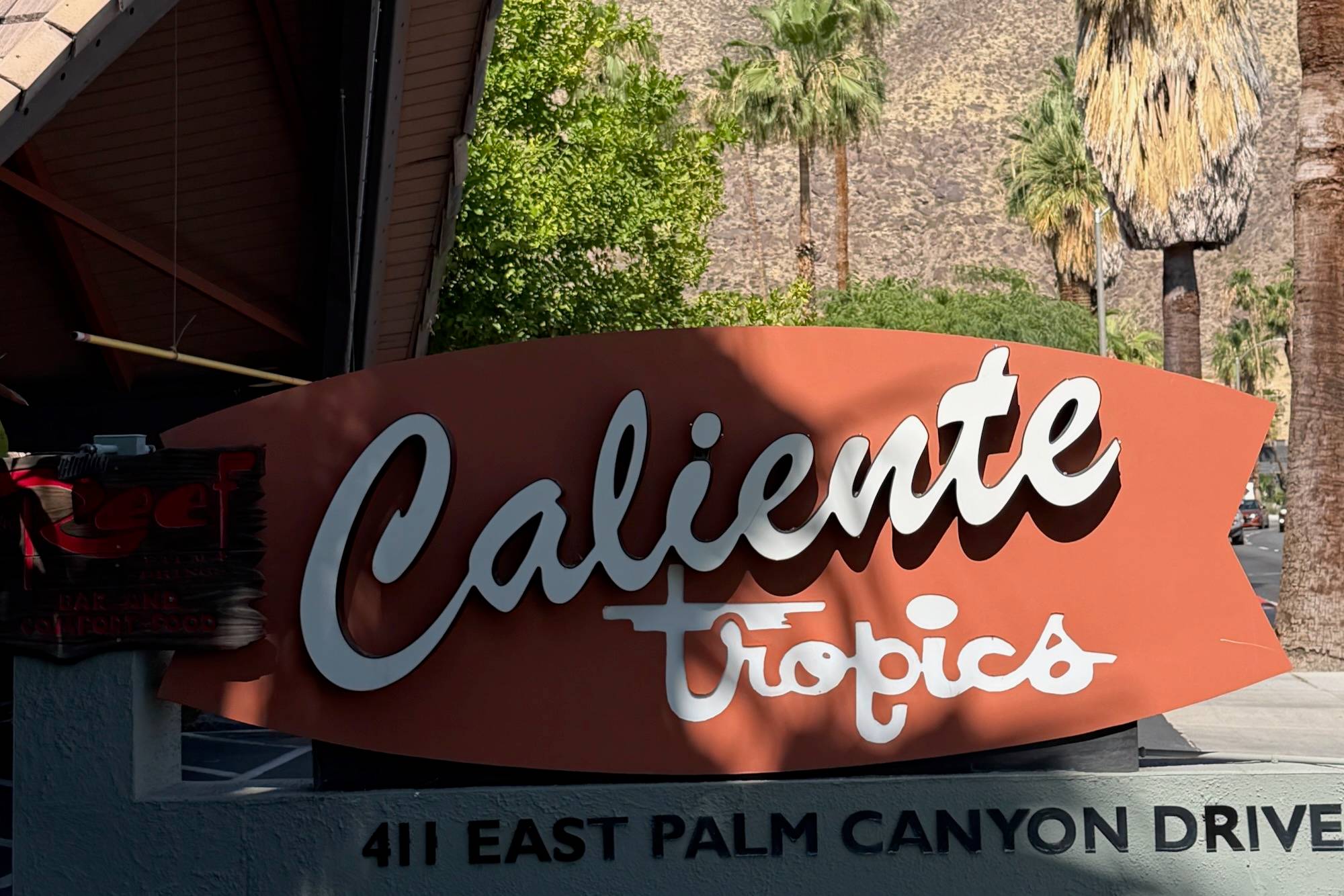 A surfboard-shaped sign reading Caliente Tropics against a blue sky.