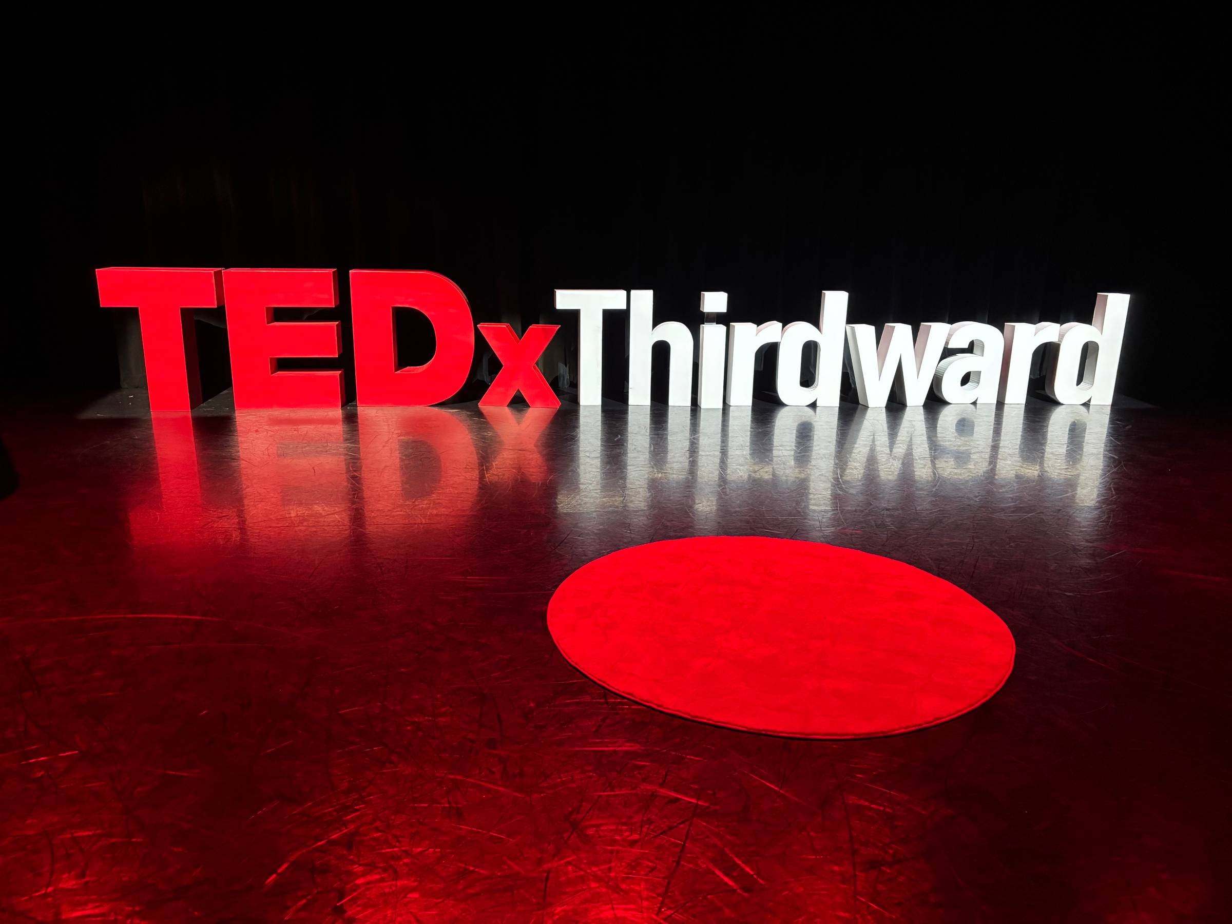 TEDx Third Ward stage with red TEDx circle and event signage before speaker presentation.